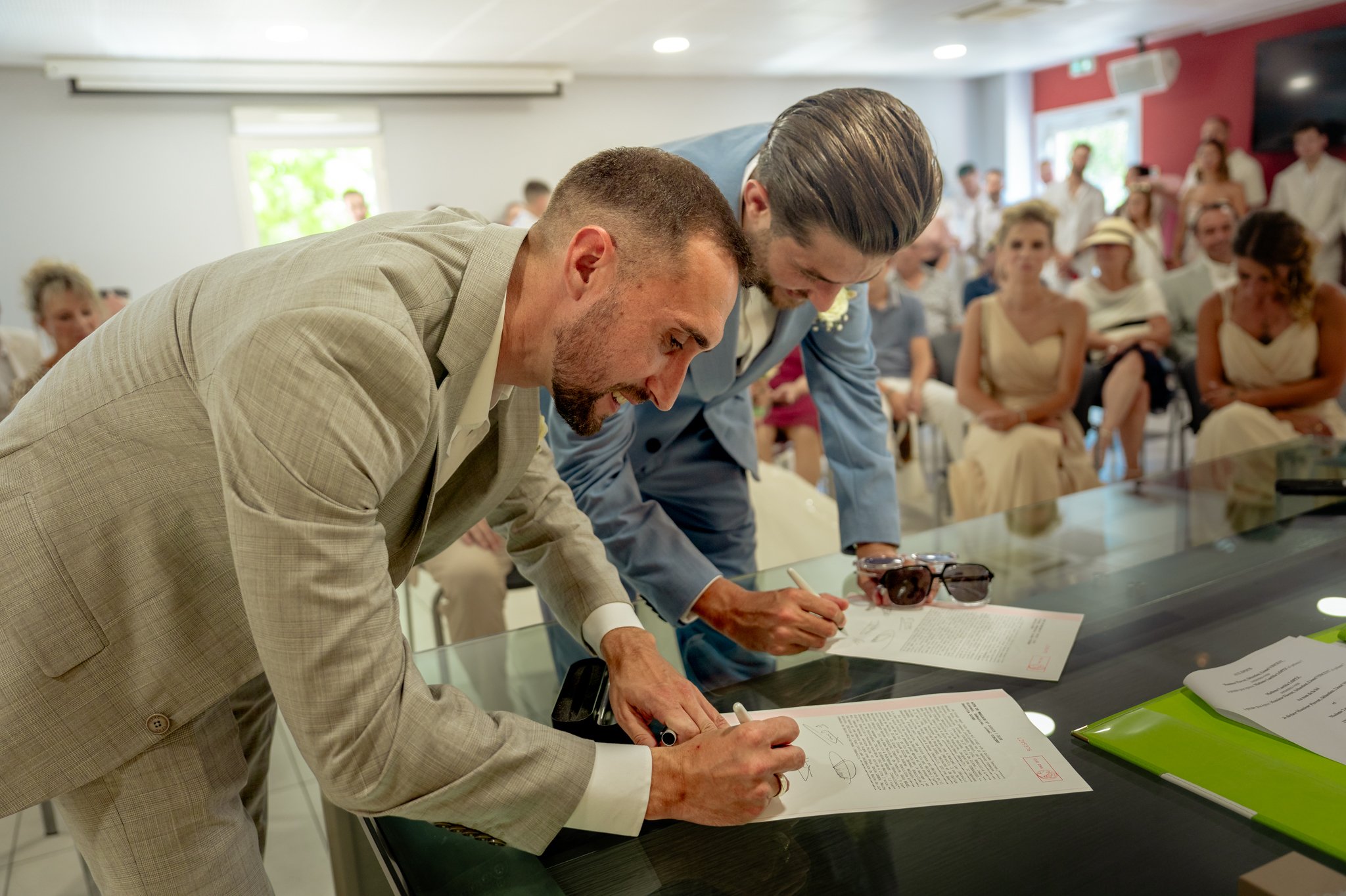 Deux hommes signent un document lors d'une cérémonie, entourés de nombreux invités assis, certains prennent des photos.