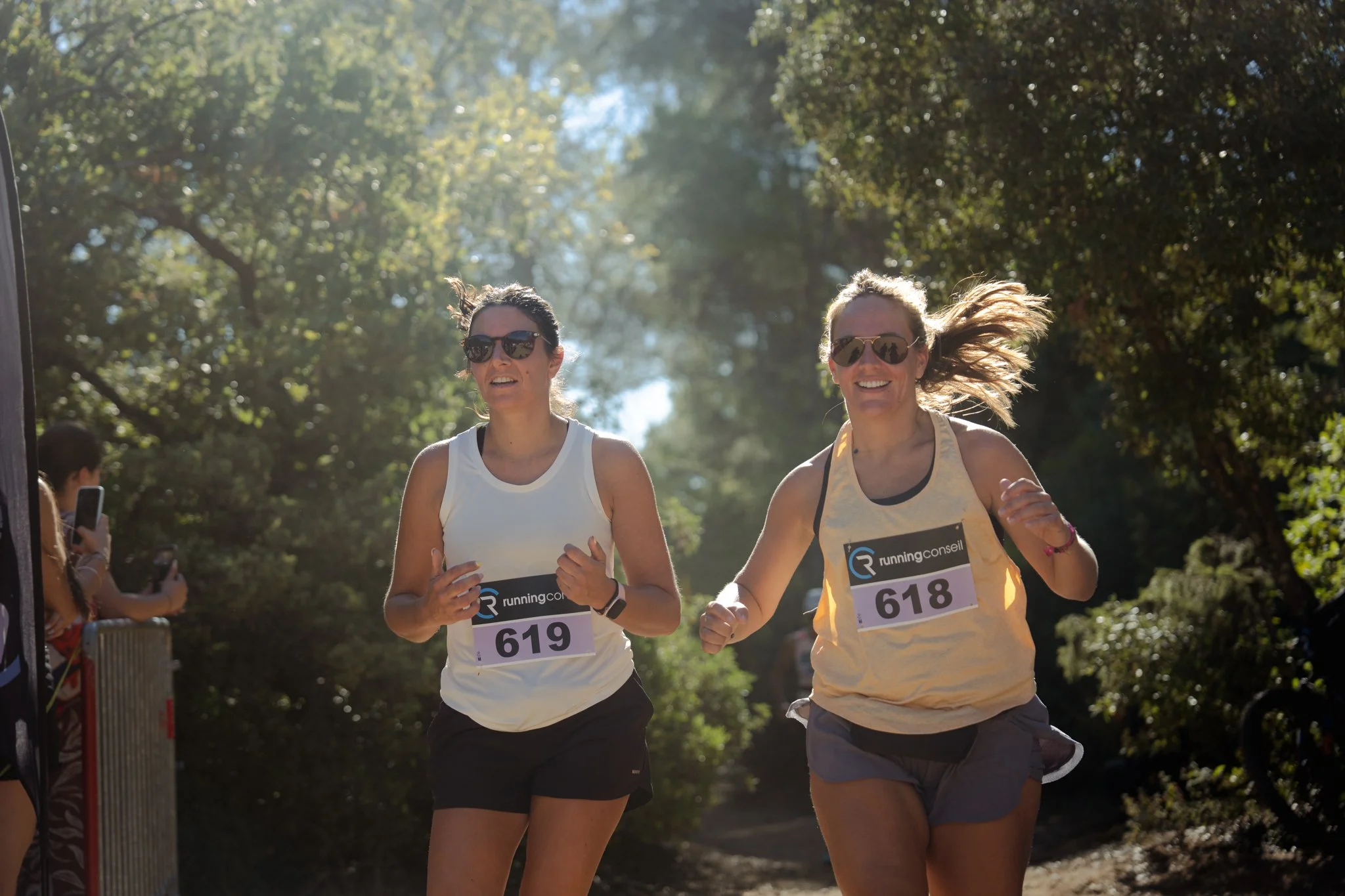 Deux femmes courent lors d'une course en plein air, portant des numéros 618 et 619 sur leur t-shirt, sous un ciel ensoleillé et entourées d'arbres.