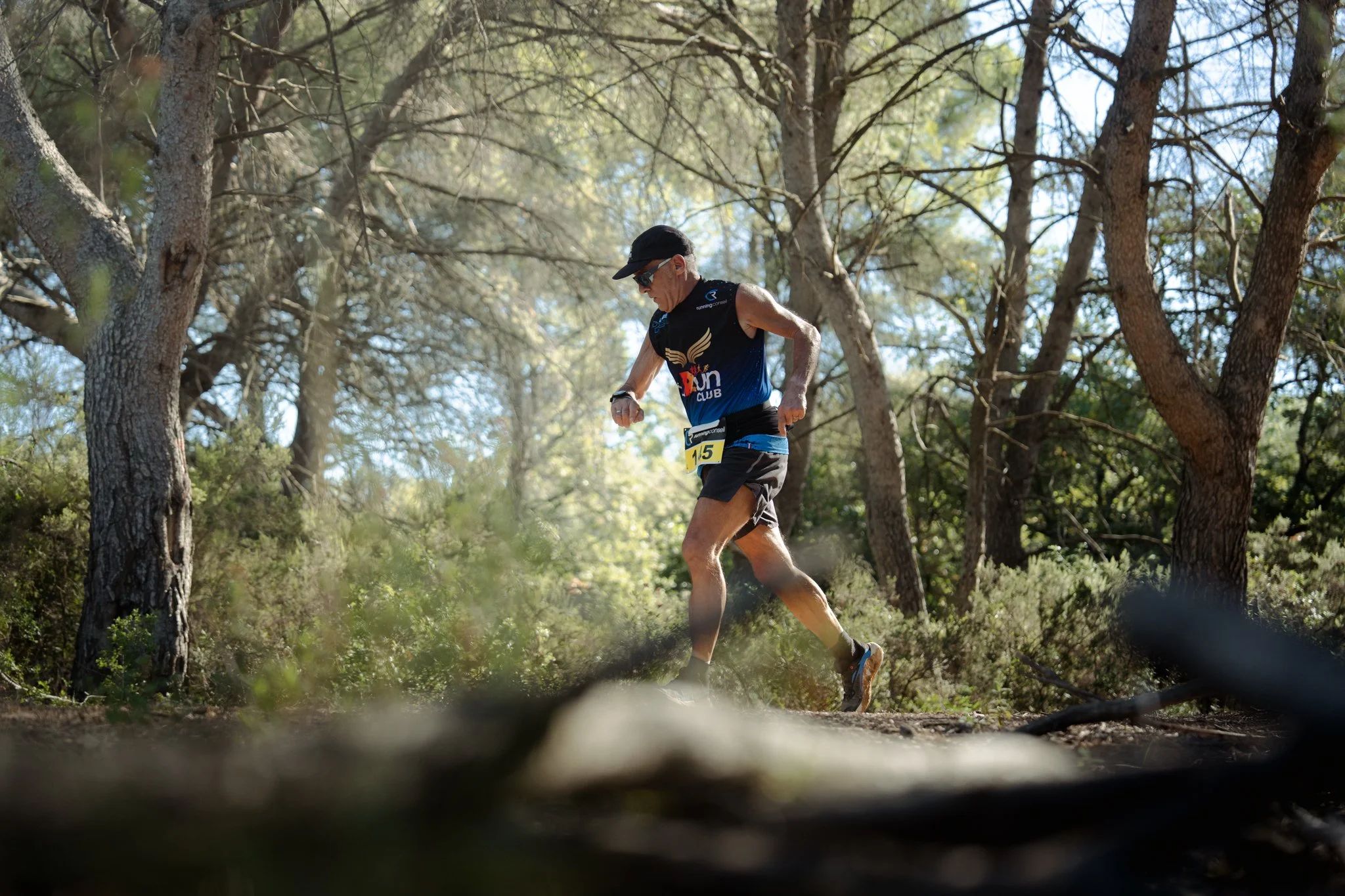 Un homme court dans une forêt, portant un casque de course, des lunettes de soleil, un maillot sans manches et un short, avec un numéro de course attaché à la taille.