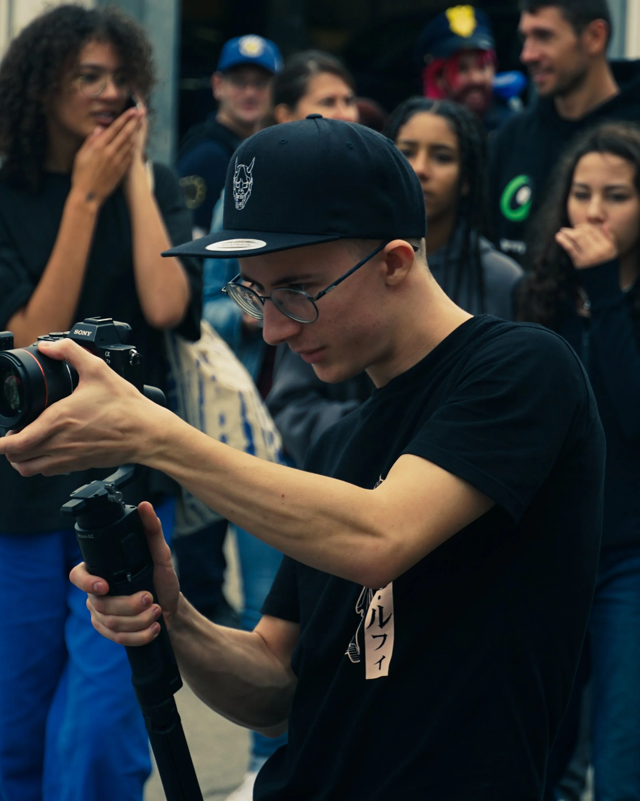 Un groupe de jeunes personnes en plein air, dont un jeune homme porte une casquette noire avec un logo de démon, manipulant un trépied ou un support pour une caméra ou un appareil photo. D'autres personnes autour regardent ou partagent l'instant, vêtues de vêtements décontractés et certains avec des accessoires comme des casques ou des écouteurs.