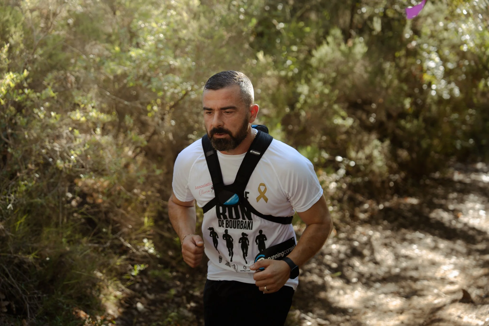 Un homme courant dans un sentier forestier, portant un t-shirt blanc, un sac à dos, et une montre connectée.