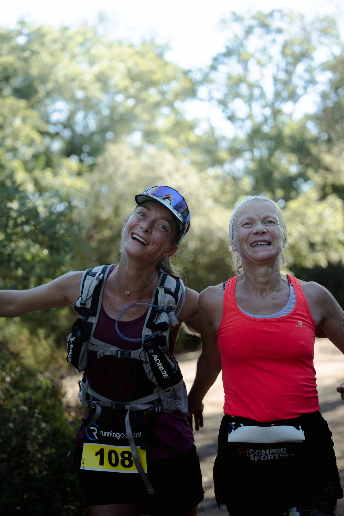 Deux femmes courent en plein air dans un environnement naturel entouré d'arbres, l'une porte un casque de vélo et un gilet de course, et l'autre porte un débardeur rouge. Elles semblent heureuses et en pleine forme.
