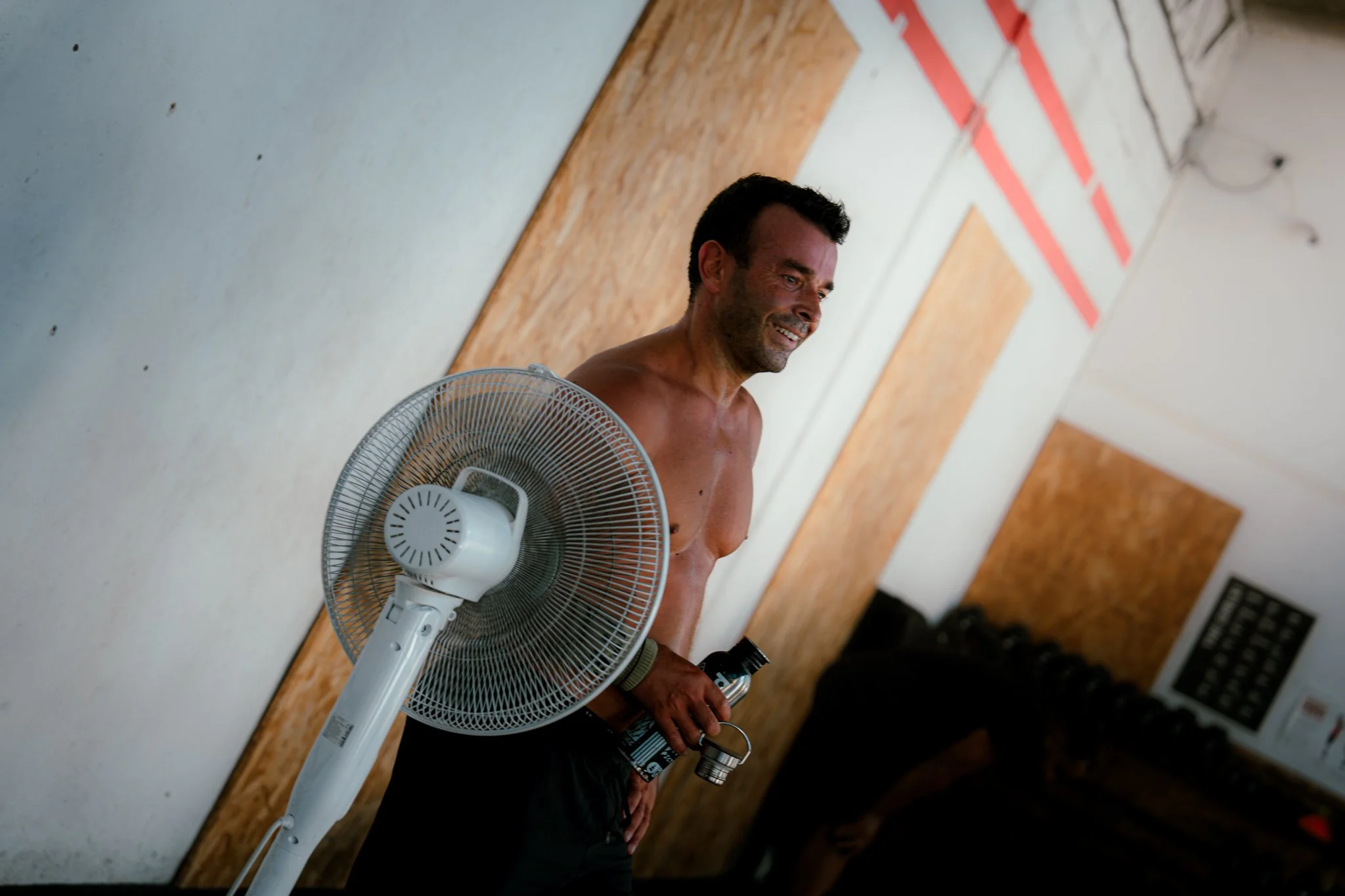 Un homme torse nu se tenant près d'un ventilateur de table dans une pièce avec un mur en bois et des câbles électriques au plafond.