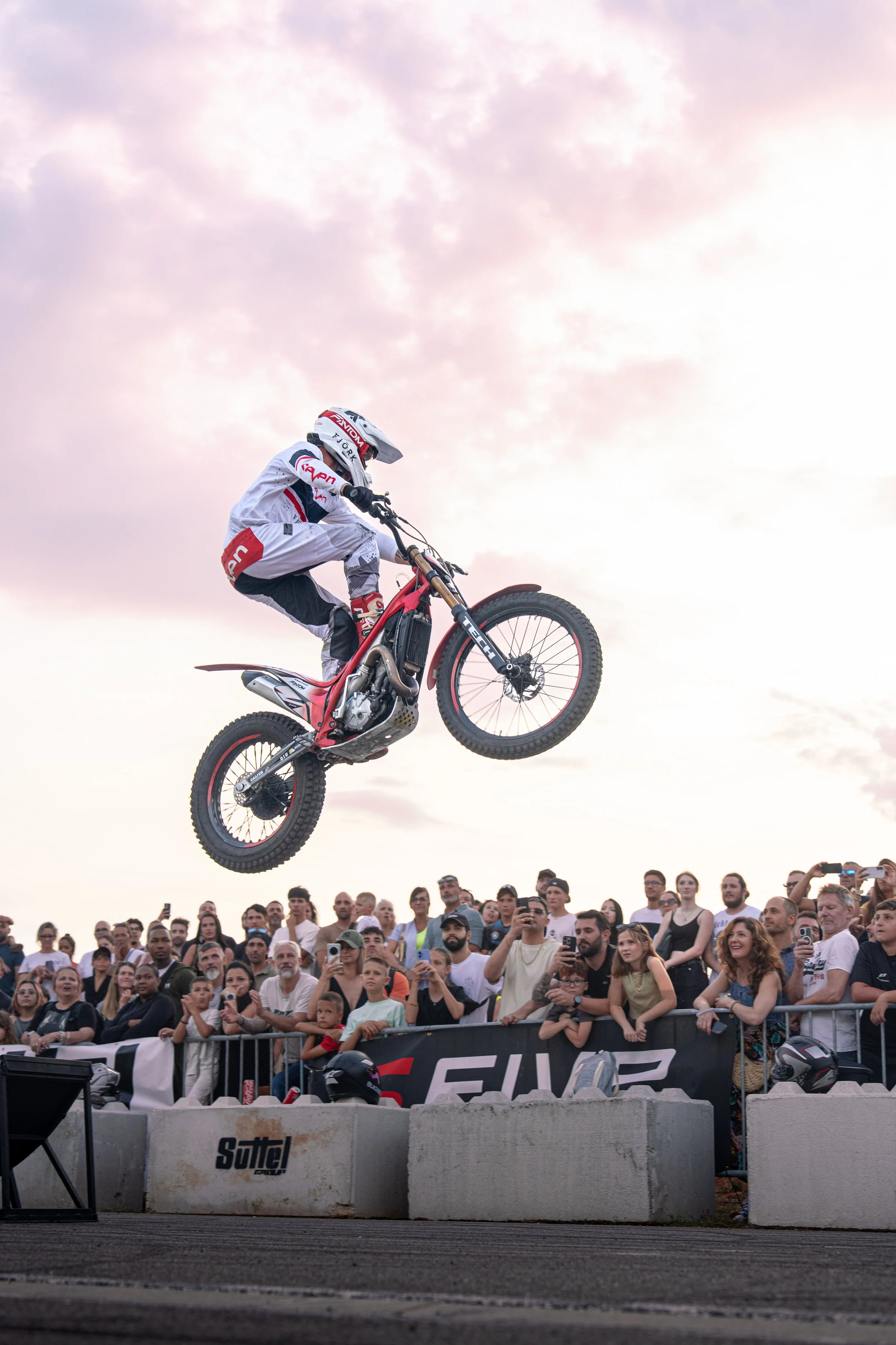Motocross rider performing a jump in front of a crowd of spectators at an outdoor event during sunset.