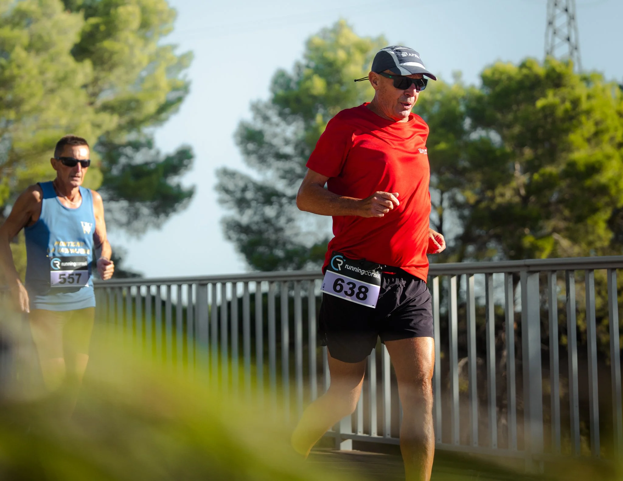 Deux coureurs, hommes âgés, participent à une course en extérieur, portant des vêtements de course et des numéros de dossard, avec un environnement naturel et un ciel clair en arrière-plan.