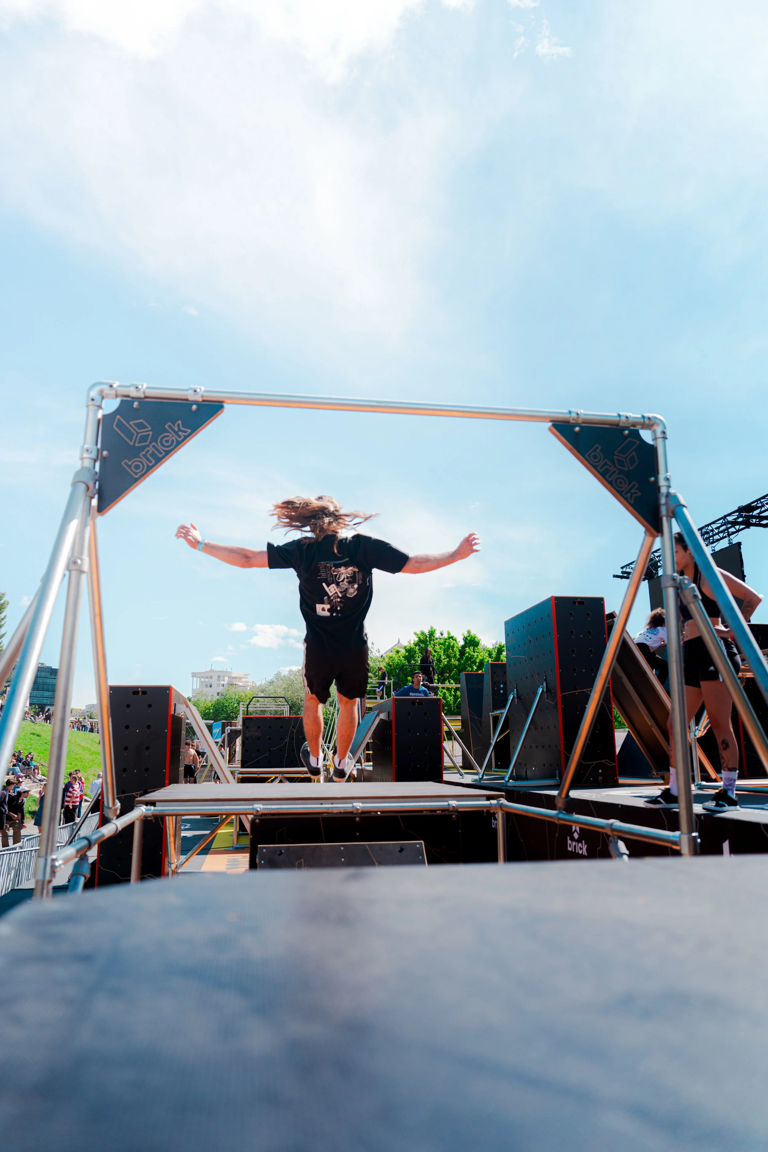 Un homme en train de sauter sur un trampoline lors d'un événement en plein air sous un ciel bleu avec quelques nuages.