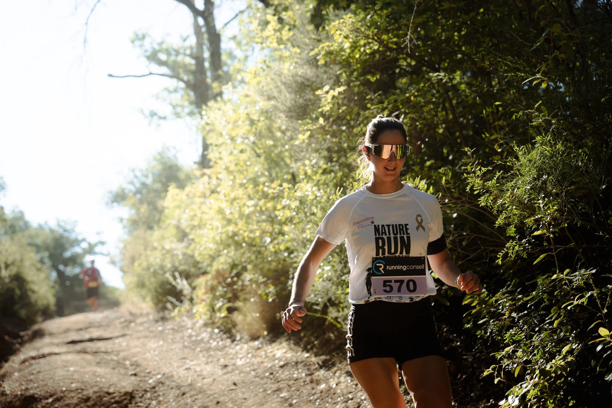 Femme courant dans un sentier forestier en pleine nature avec lunettes de soleil et dossard de course.