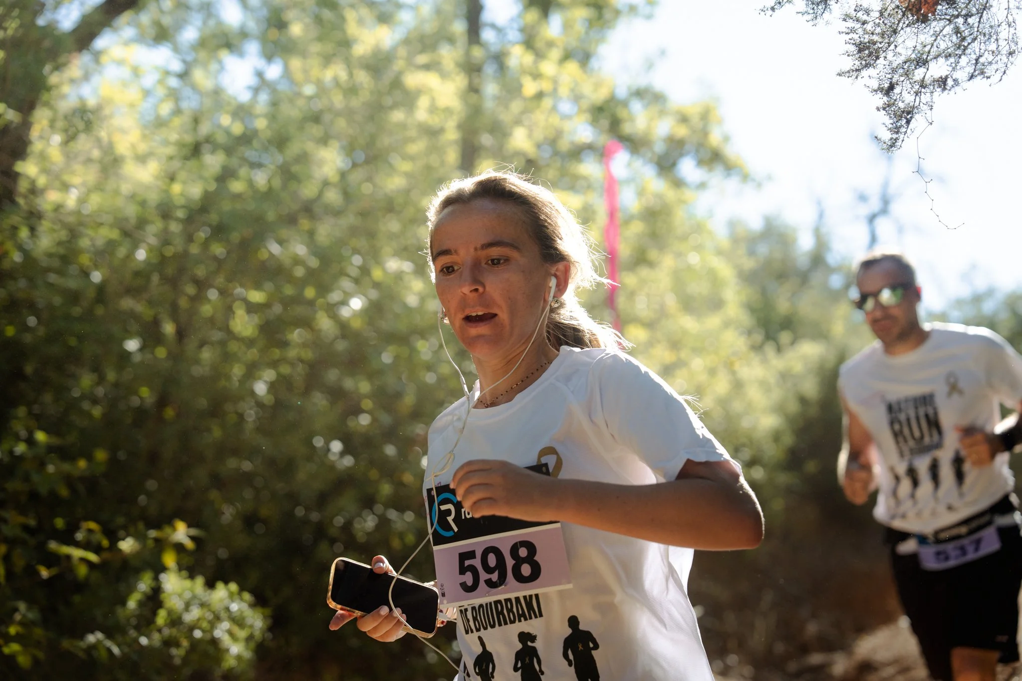 Deux coureurs participent à une course dans un espace naturel, portant des dossards avec des numéros. La femme en premier plan porte des écouteurs et regarde son téléphone, tandis qu'un homme la suit juste derrière. La scène est en plein air, avec be