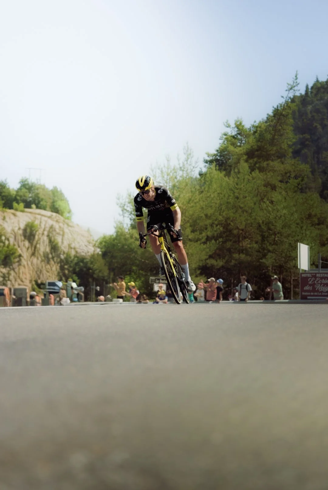 A cyclist wearing a black and yellow uniform and helmet rides a bike on a rural road with green trees and a crowd of spectators in the background.