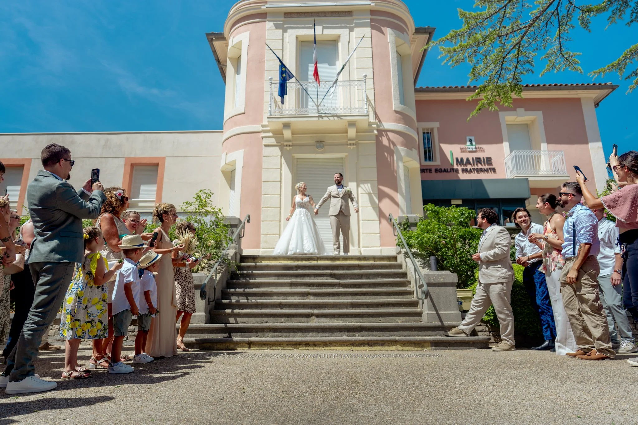 Un groupe de personnes assiste à un mariage devant une mairie. La mariée en robe blanche et le marié en costume marchent côte à côte. La mairie est une bâtisse rose claire avec des drapeaux français et européen suspendus au balcon. Les invités, habillés en tenues légères, prennent des photos et regardent la cérémonie.