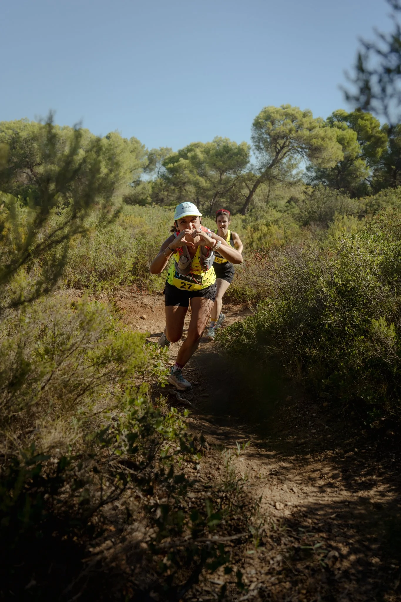 Deux femmes faisant une course à pied dans une forêt ensoleillée, portant des tenues de sport jaunes avec des numéros de course.