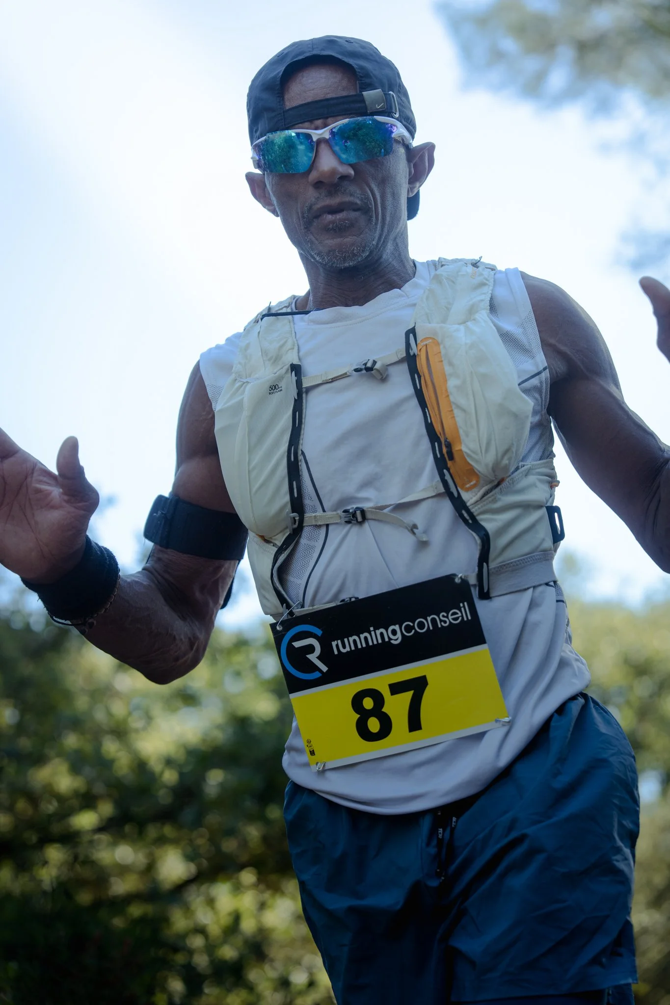 Un homme courant lors d'une compétition de course à pied en plein air, portant un numéro de racer 87, avec des lunettes de soleil, un chapeau noir et une tenue de sport légère, entouré d'arbres.