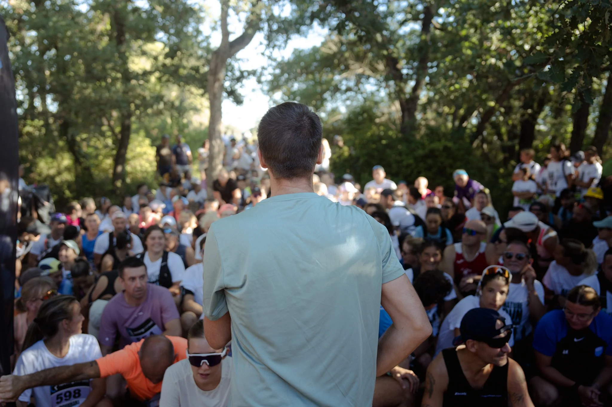 Un homme vu de dos, par-derrière, semble parler à une grande foule lors d'un rassemblement en plein air dans un parc, entouré de nombreux spectateurs.