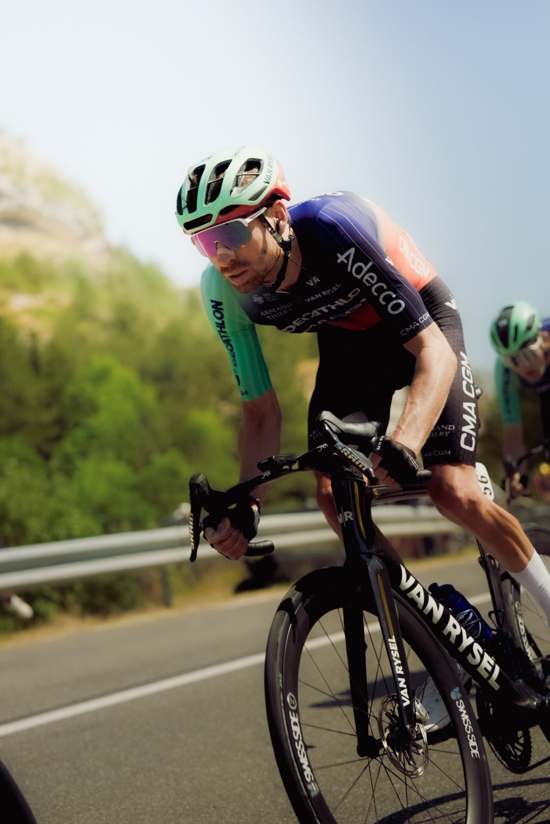 A male cyclist in a racing uniform and helmet riding on a mountain road, with greenery and a guardrail in the background.