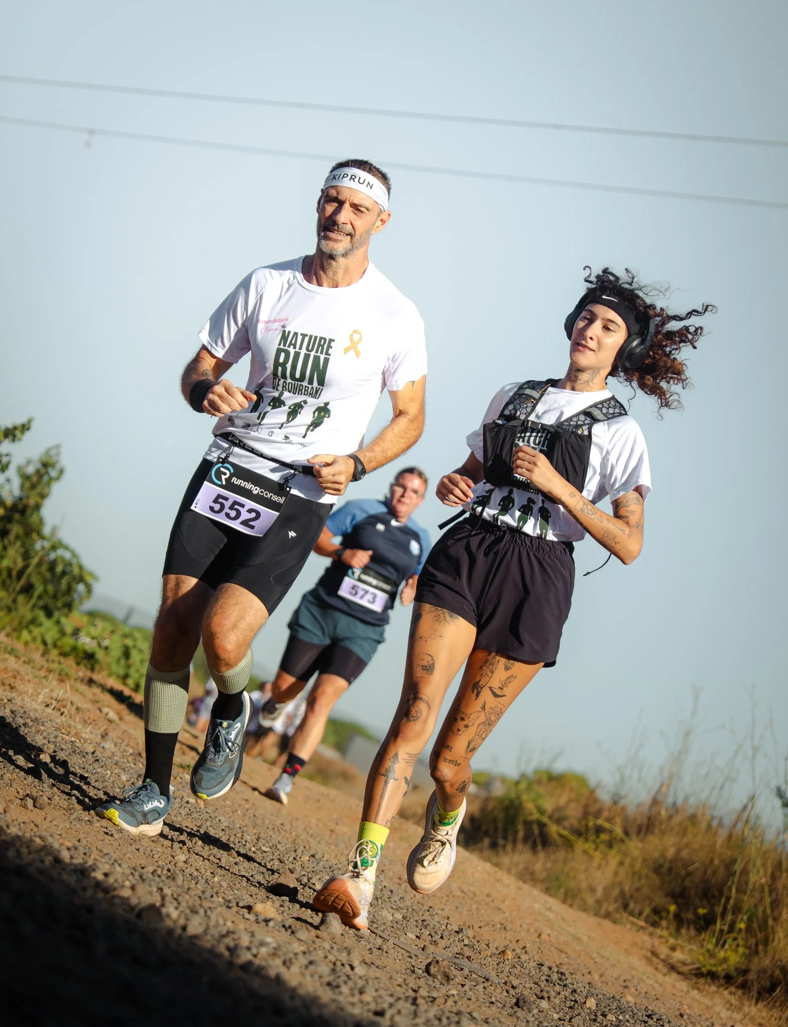 Groupe de coureurs participant à une course sur un sentier en plein air, certains sont tatoués et portent des vêtements de sport, avec un ciel clair en arrière-plan.