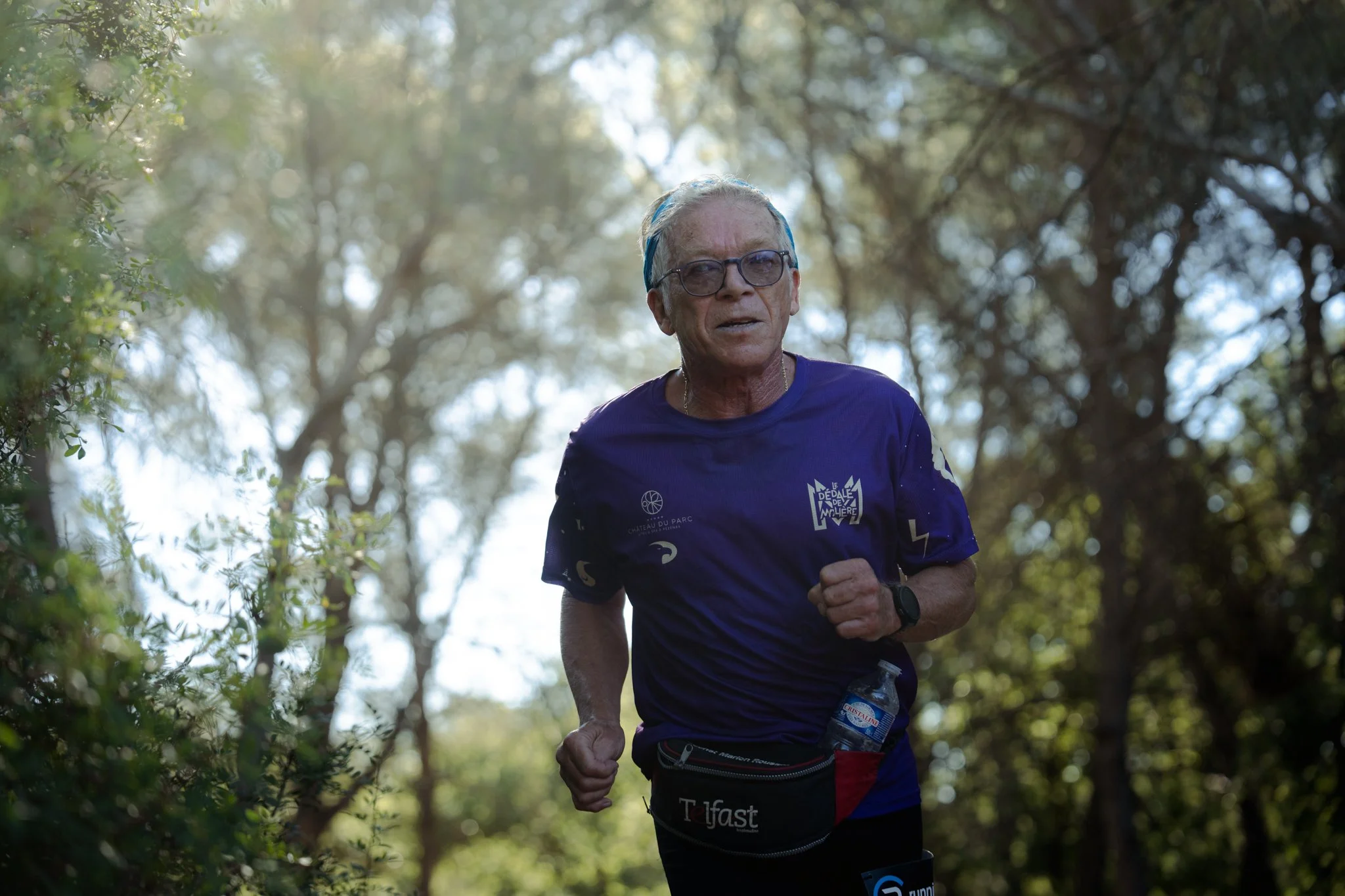 Un homme âgé court dans un sentier forestier, portant une tenue de course violette avec des logos, une montre, une bouteille d'eau attachée à la taille, et des lunettes.
