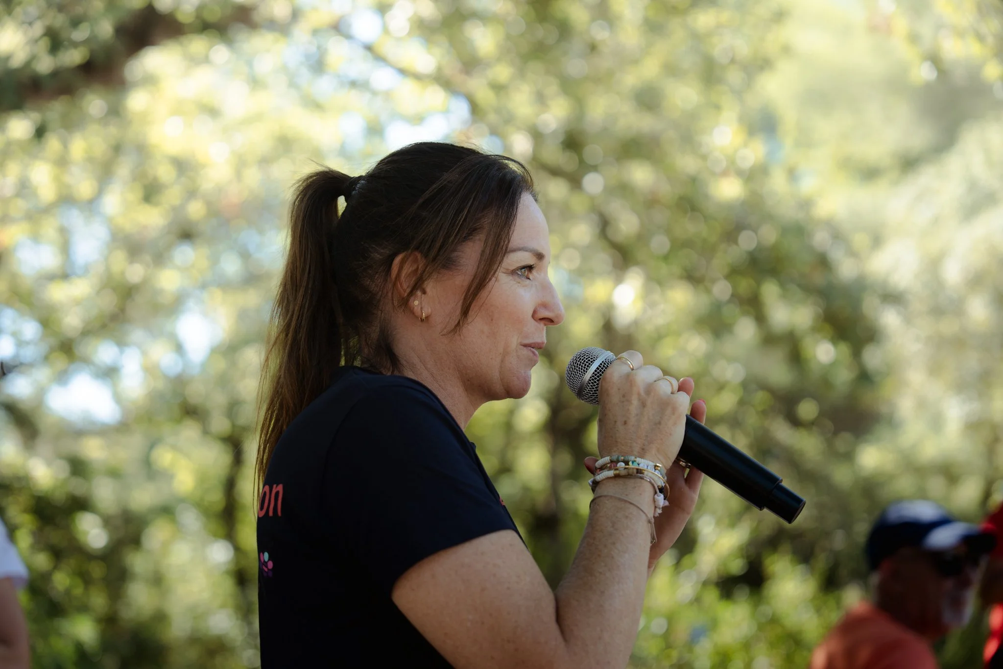 Une femme parle dans un microphone lors d'un événement en plein air, avec un fond de feuilles d'arbres ensoleillées.