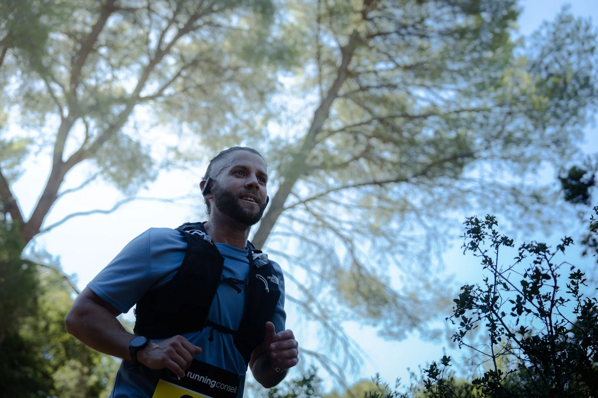 Homme courant dans une forêt verdoyante, portant un casque de course, un sac à dos, une montre et un t-shirt bleu, avec un ciel bleu et des arbres feuillus en arrière-plan.
