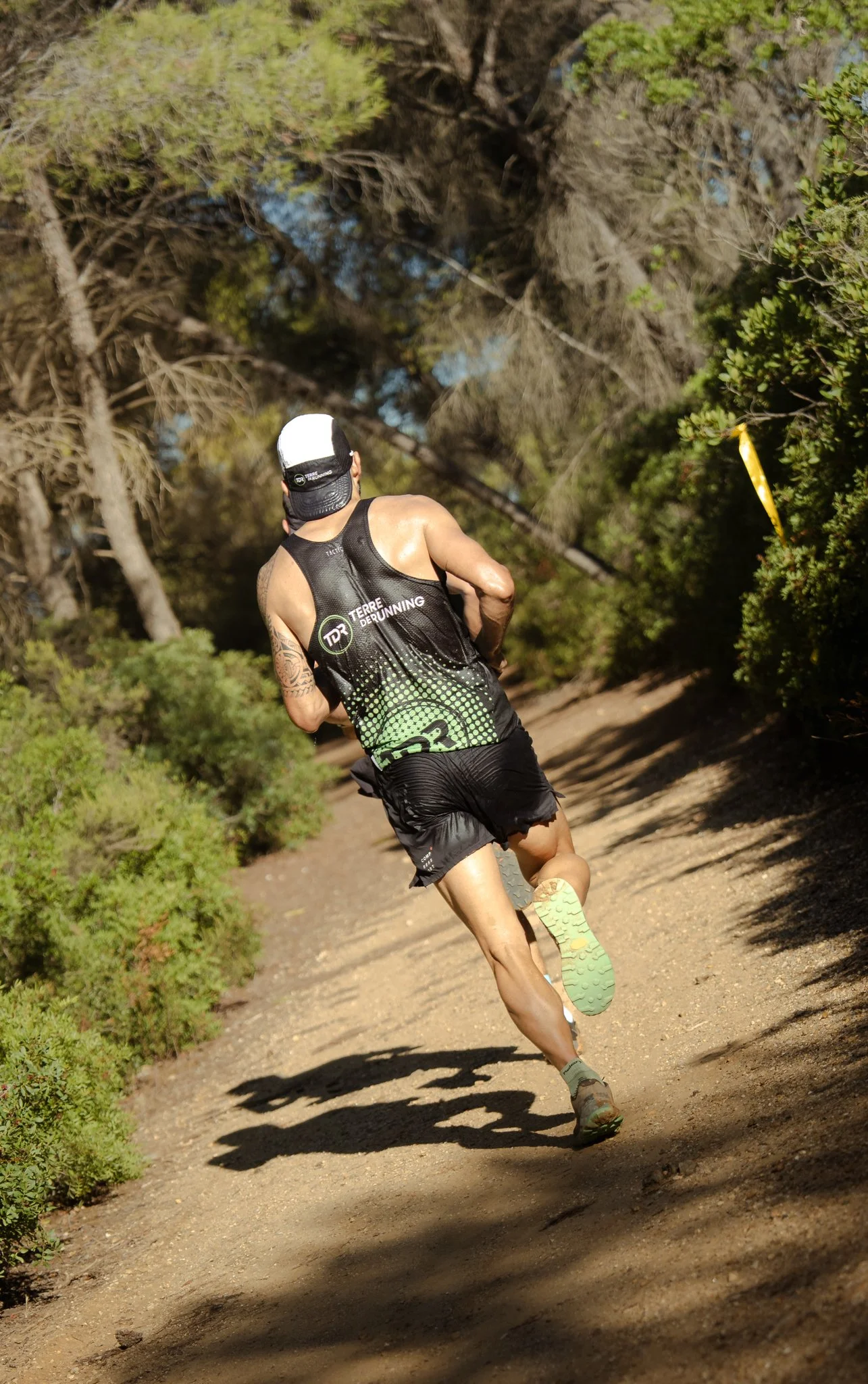 Un homme habillé en course à pied sur un sentier de forêt, portant un maillot noir avec le texte 'Terre De Running' et un chapeau blanc et noir, entouré d'arbres et de buissons.