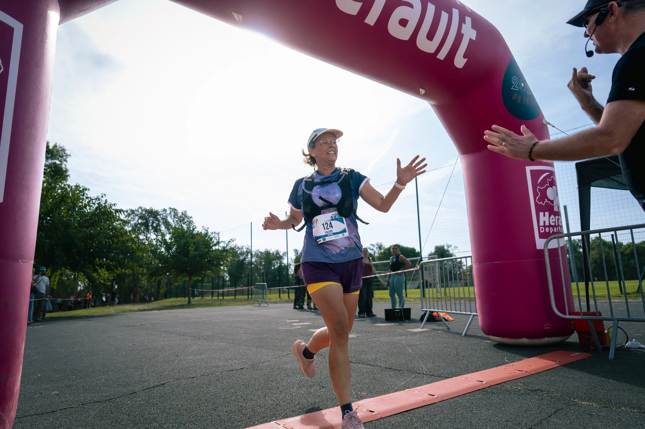 Une femme court la ligne d'arrivée de la course, franchissant la zone de finition sous un arc gonflable rosa avec des textes en français, portant un dossard numéro 124, un chapeau, des lunettes et une tenue de sport colorée, tandis que d'autres participants et spectateurs se trouvent en arrière-plan.