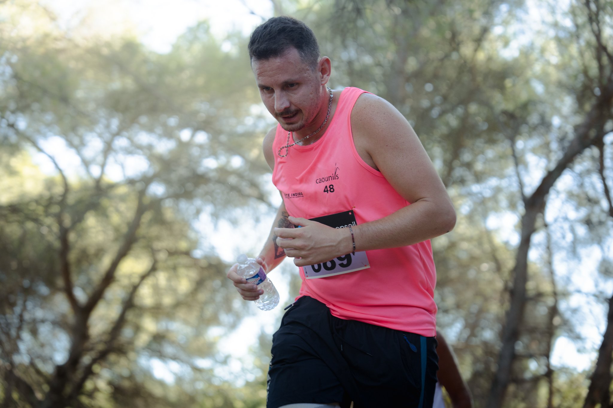 Un homme court dans un parc en portant un maillot rose avec un numéro de course, il tient une bouteille d'eau dans la main.