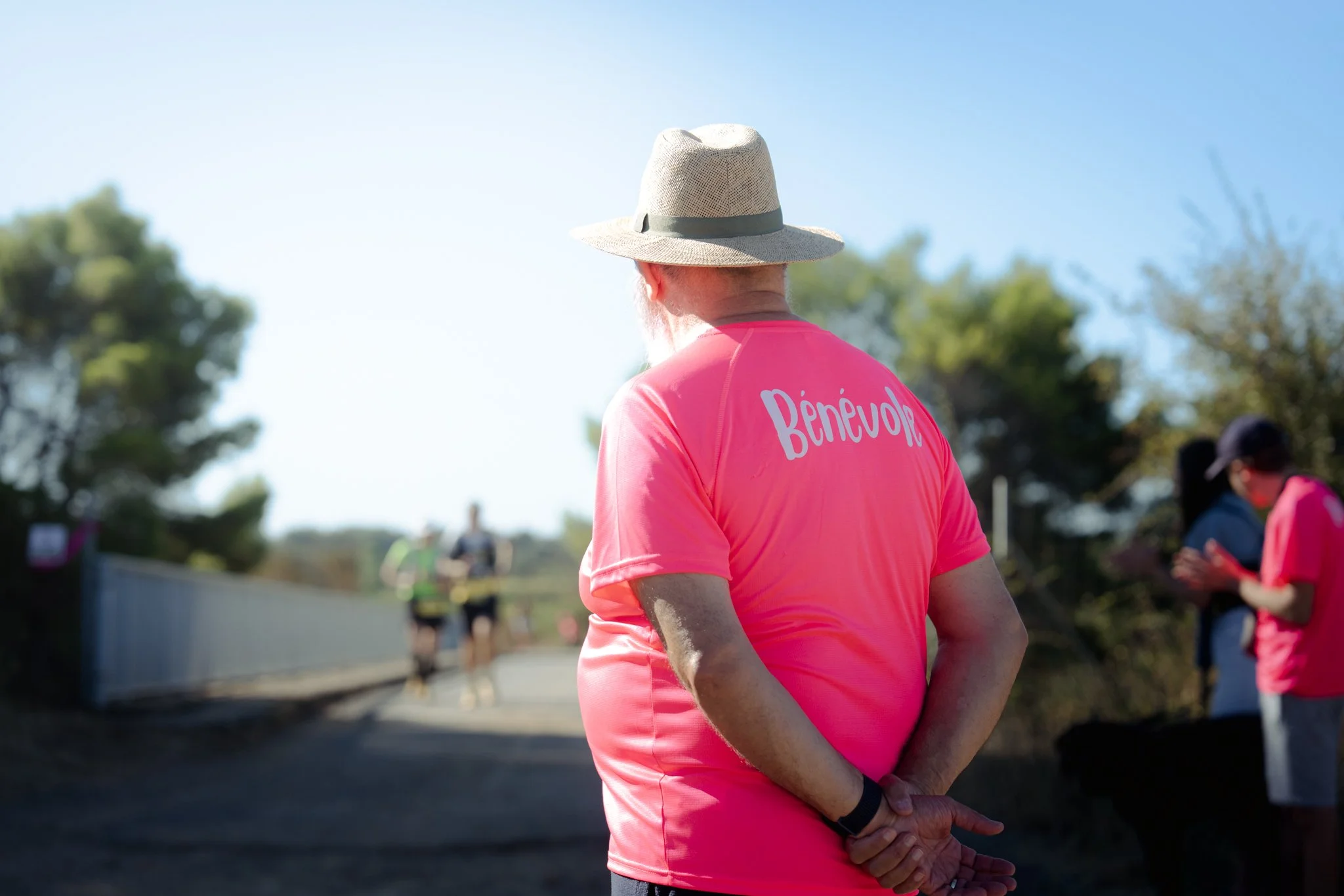 Un homme âgé portant un chapeau beige et un t-shirt rose avec l'inscription 'Bénévol' regardant vers l'horizon lors d'un événement en plein air, avec d'autres personnes en arrière-plan sous un ciel clair.