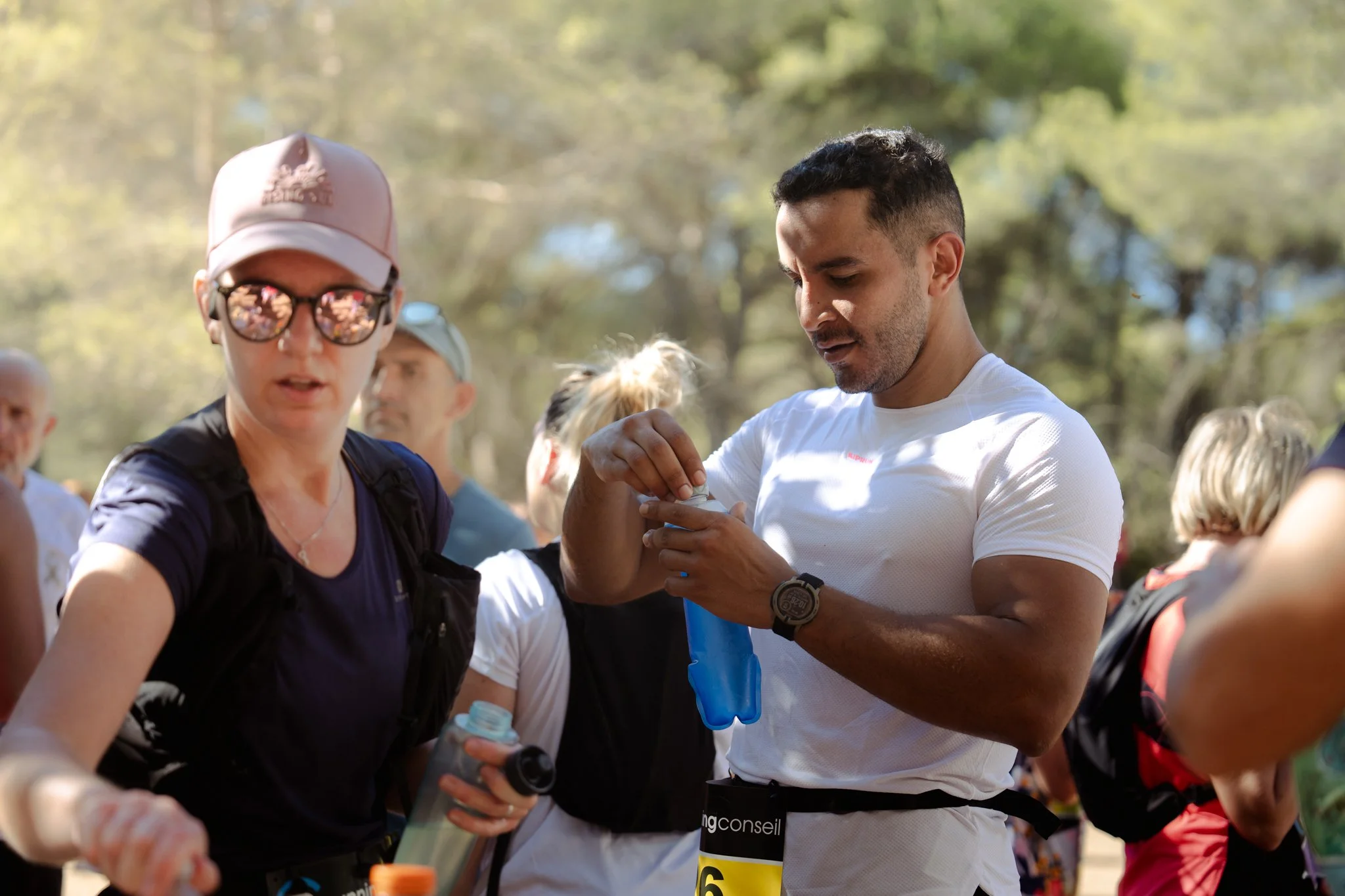 Groupe de personnes participant à une course ou un événement sportif en plein air, avec un homme en t-shirt blanc et une femme portant une casquette et des lunettes de soleil, tous deux préparant leur équipement.