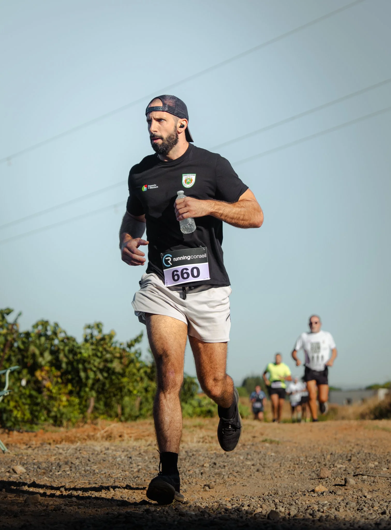 Homme courant lors d'une course en plein air, portant un t-shirt noir avec un logo et un dossard 660, se tenant à côté d'autres coureurs dans un environnement naturel.