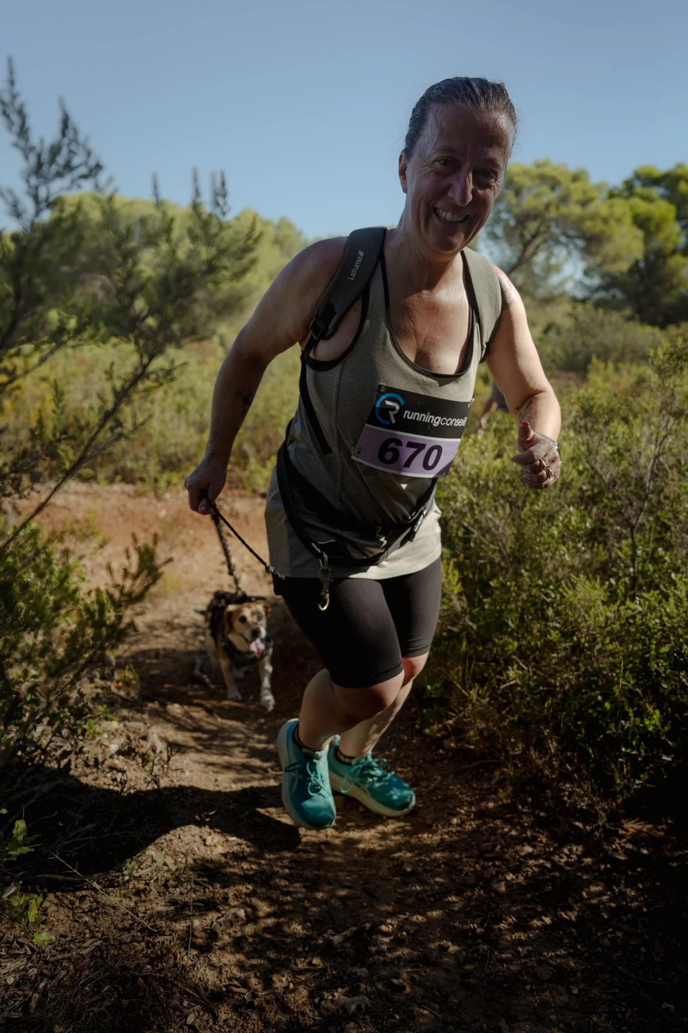 Une femme souriante court sur un sentier de nature, accompagnée de deux chiens, dans un environnement ensoleillé avec des arbres verts.