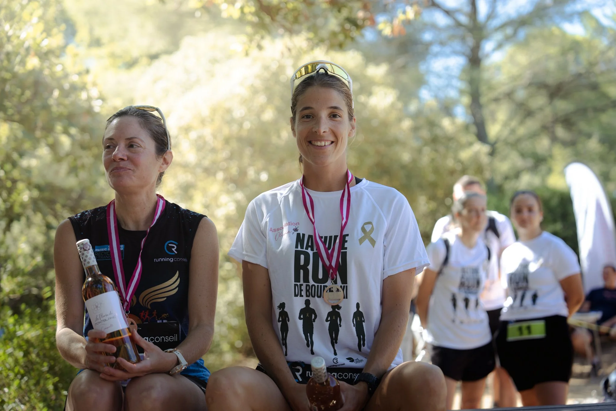 Groupe de personnes participant à une course ou une marche en extérieur, avec deux femmes en premier plan portant des médailles et des bouteilles d'eau, entourées d'arbres et de nature.