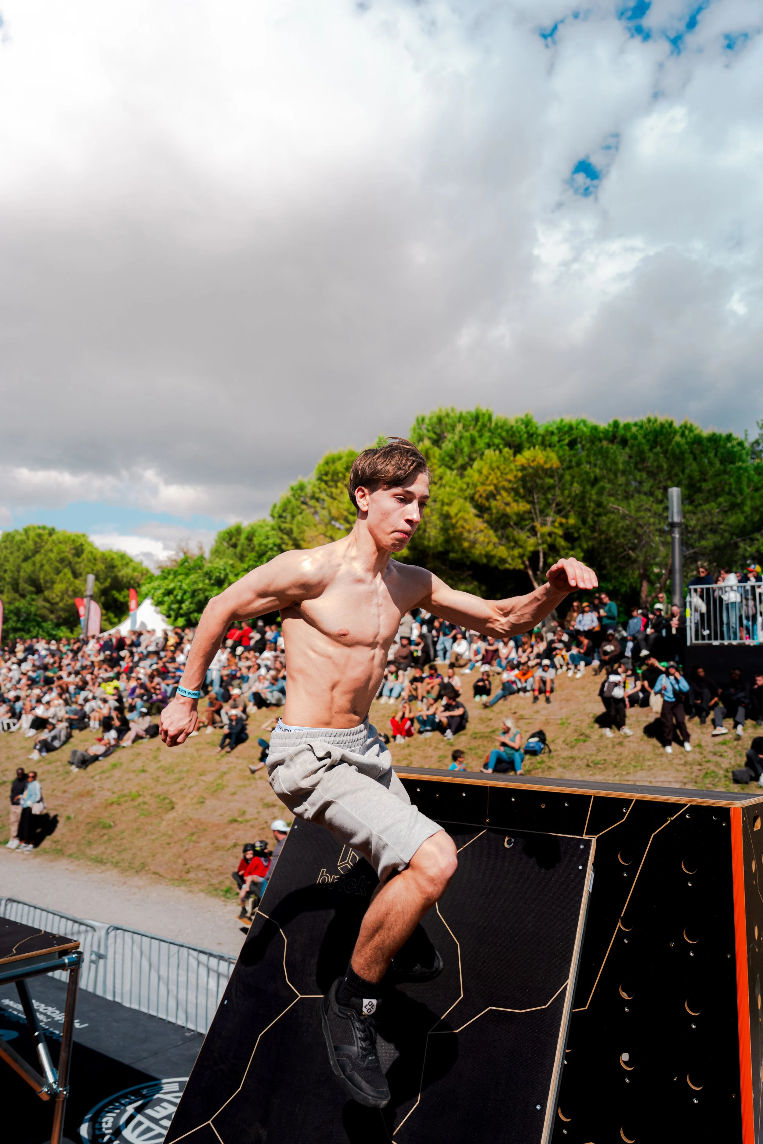 Un homme joue à la falaise lors d'une compétition de slackline en plein air, avec un public assis dans l'herbe et des arbres en arrière-plan, sous un ciel nuageux.