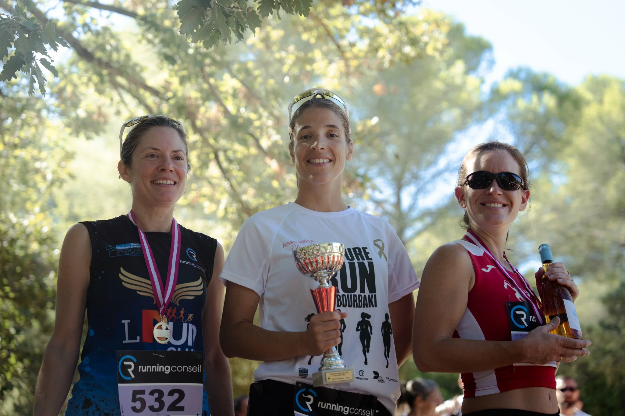 Trois femmes souriantes sur un podium lors d'une course dans un parc, portant des médailles et tenant des trophées, avec des arbres en arrière-plan.
