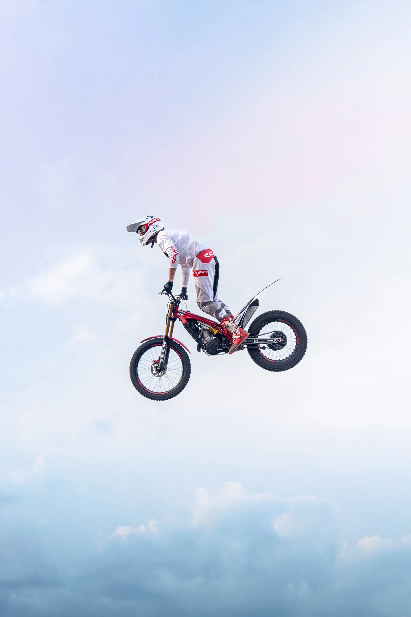 Un motard en tenue blanche effectue un saut impressionnant en moto dans le ciel avec un fond de nuages.