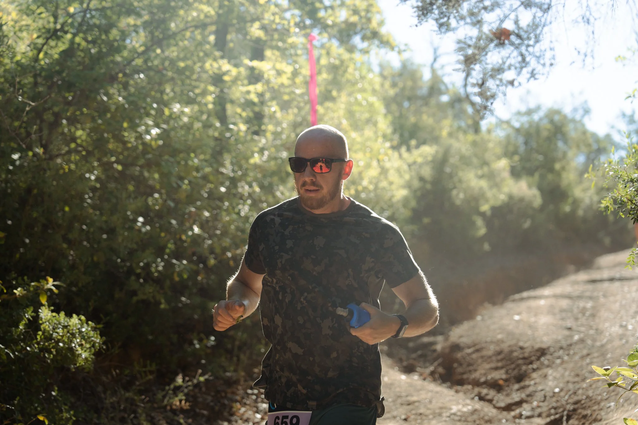 Un homme court dans une forêt ensoleillée, portant des lunettes de soleil noires, un t-shirt camouflage, un gant de course à gauche, et un dossard numéro 659.