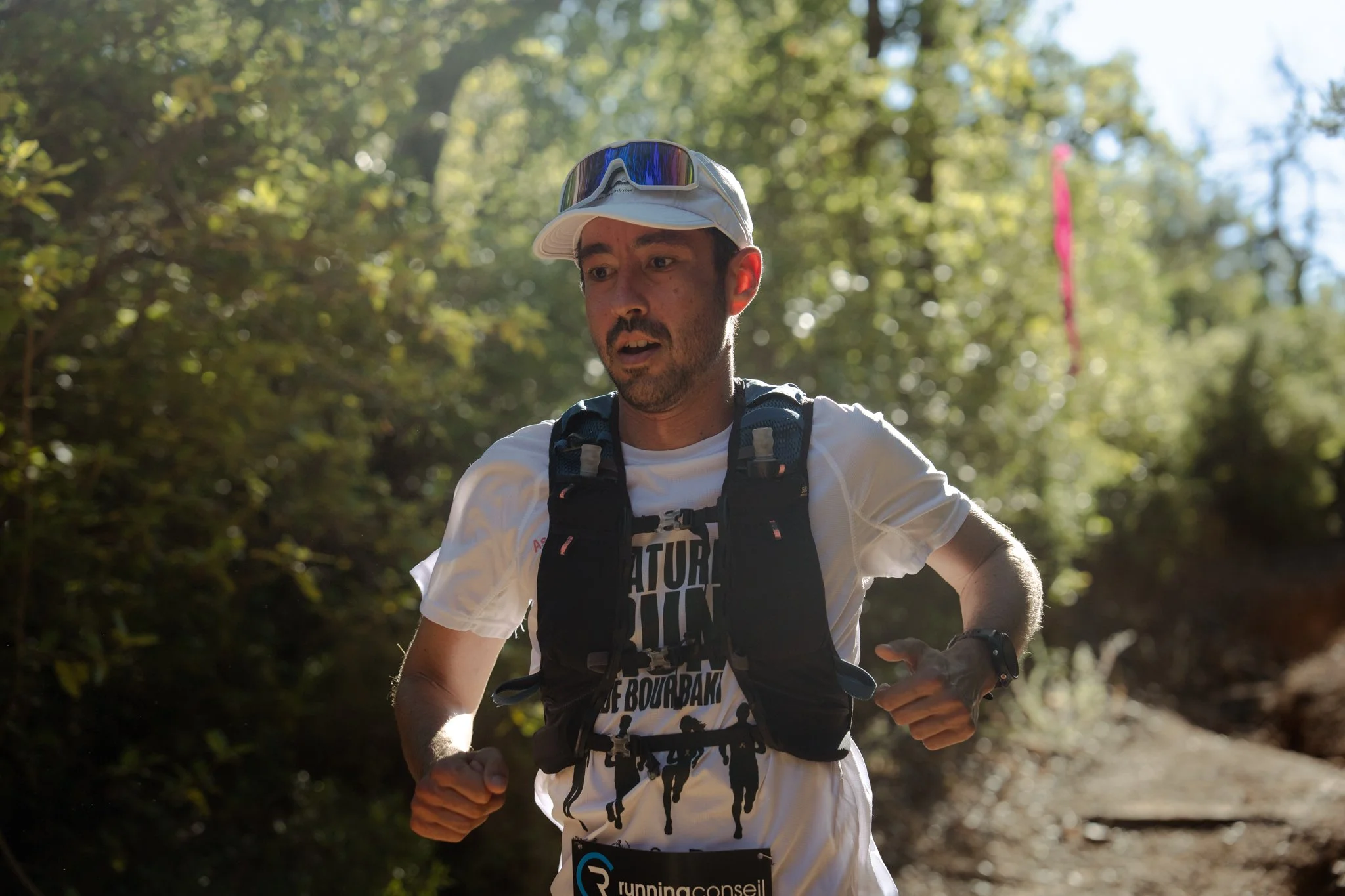 Homme courant dans une forêt verte en portant une casquette, des lunettes de soleil sur la casquette, un t-shirt blanc avec du texte, un sac à dos et une montre.