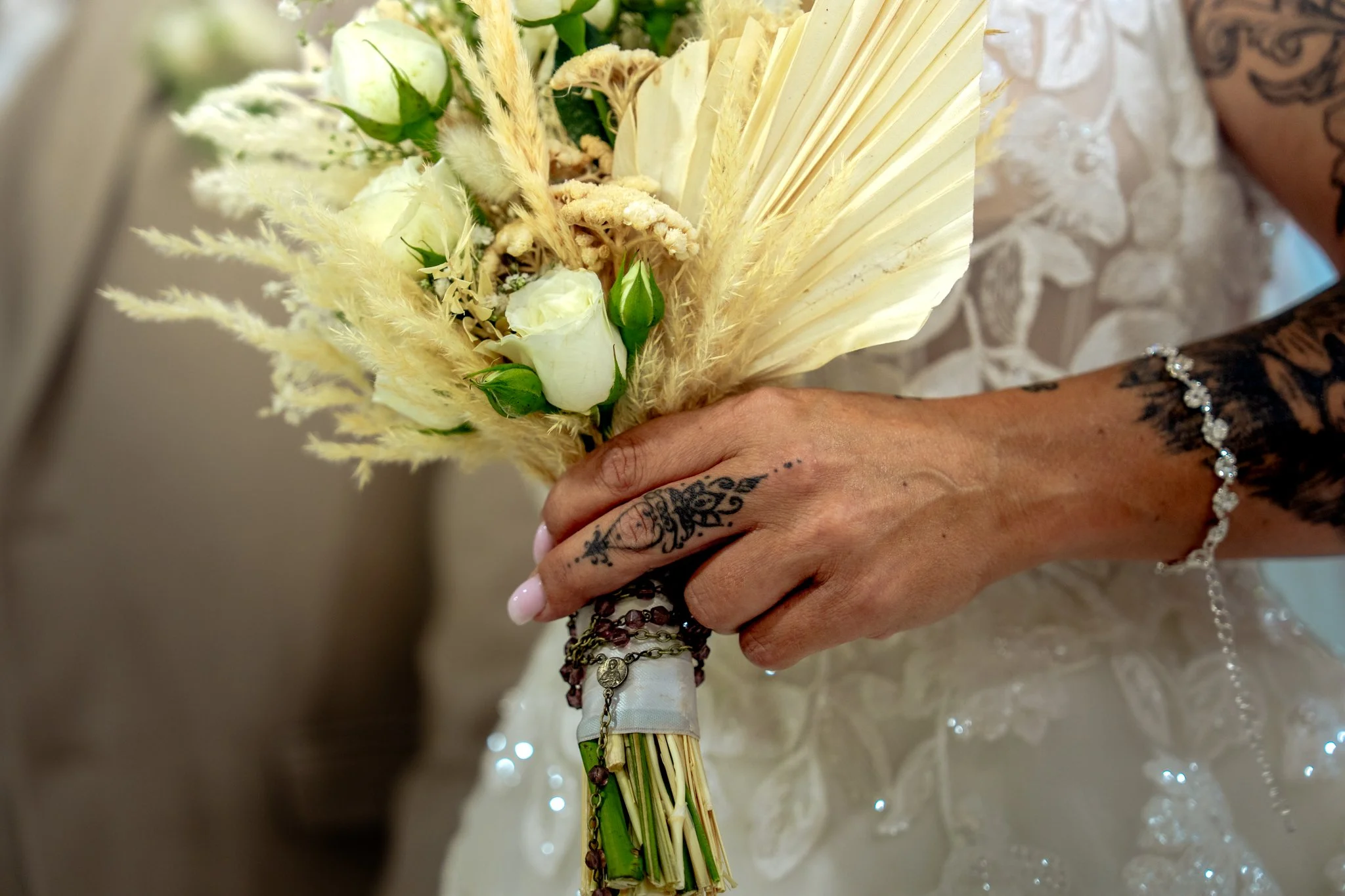 Main dans une main tatouée tenant un bouquet de fleurs blanches et de pampas, avec une robe en dentelle blanche et plusieurs bracelets.