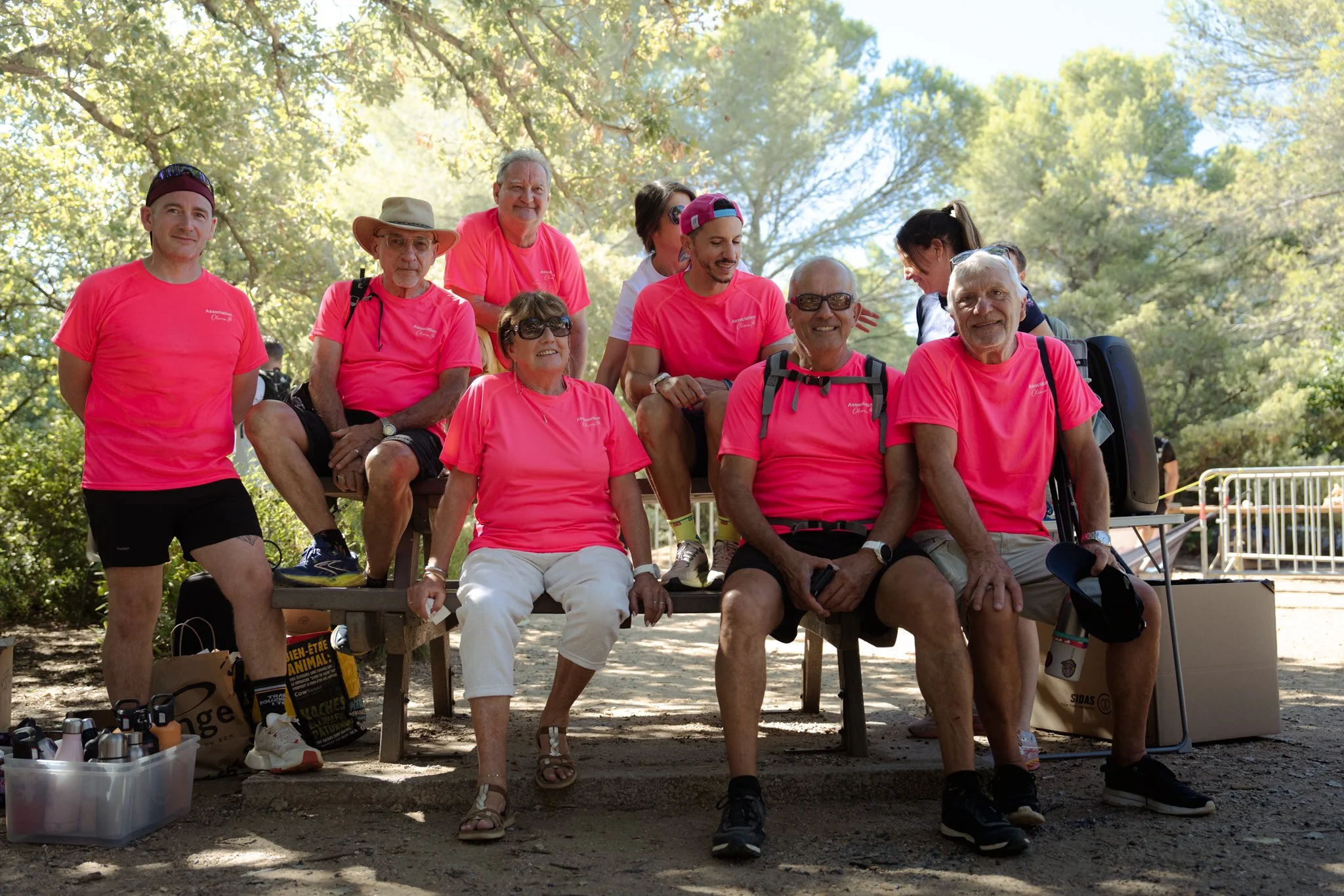 Groupe de personnes âgées et jeunes assises et debout sur un banc en plein air, portant des T-shirts roses, dans un parc avec des arbres.