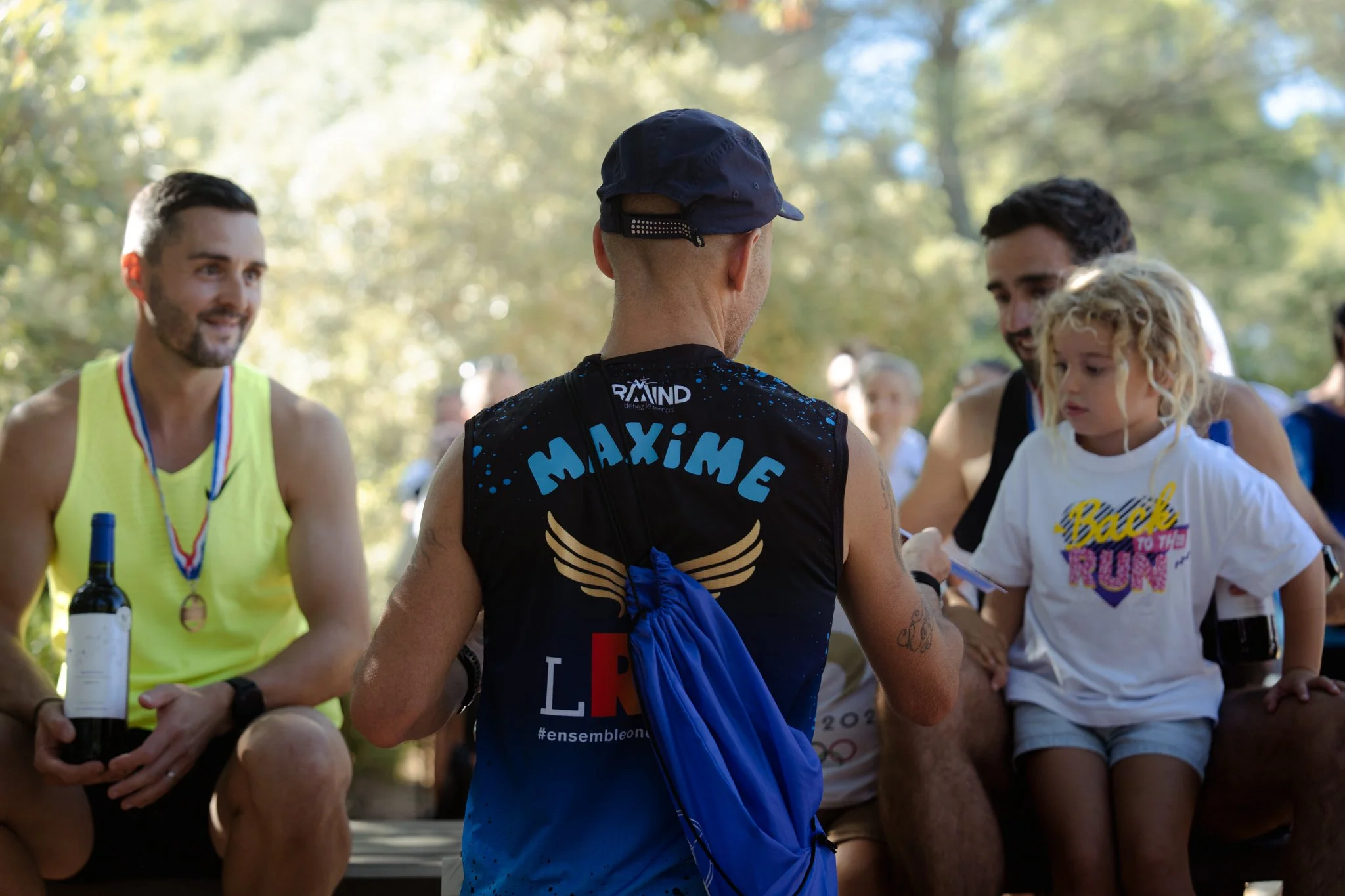 Groupe de personnes, notamment un homme portant une casquette et un maillot avec le nom Maxime, échangeant ou discutant lors d'un événement en plein air entouré de arbres, avec d'autres personnes en arrière-plan