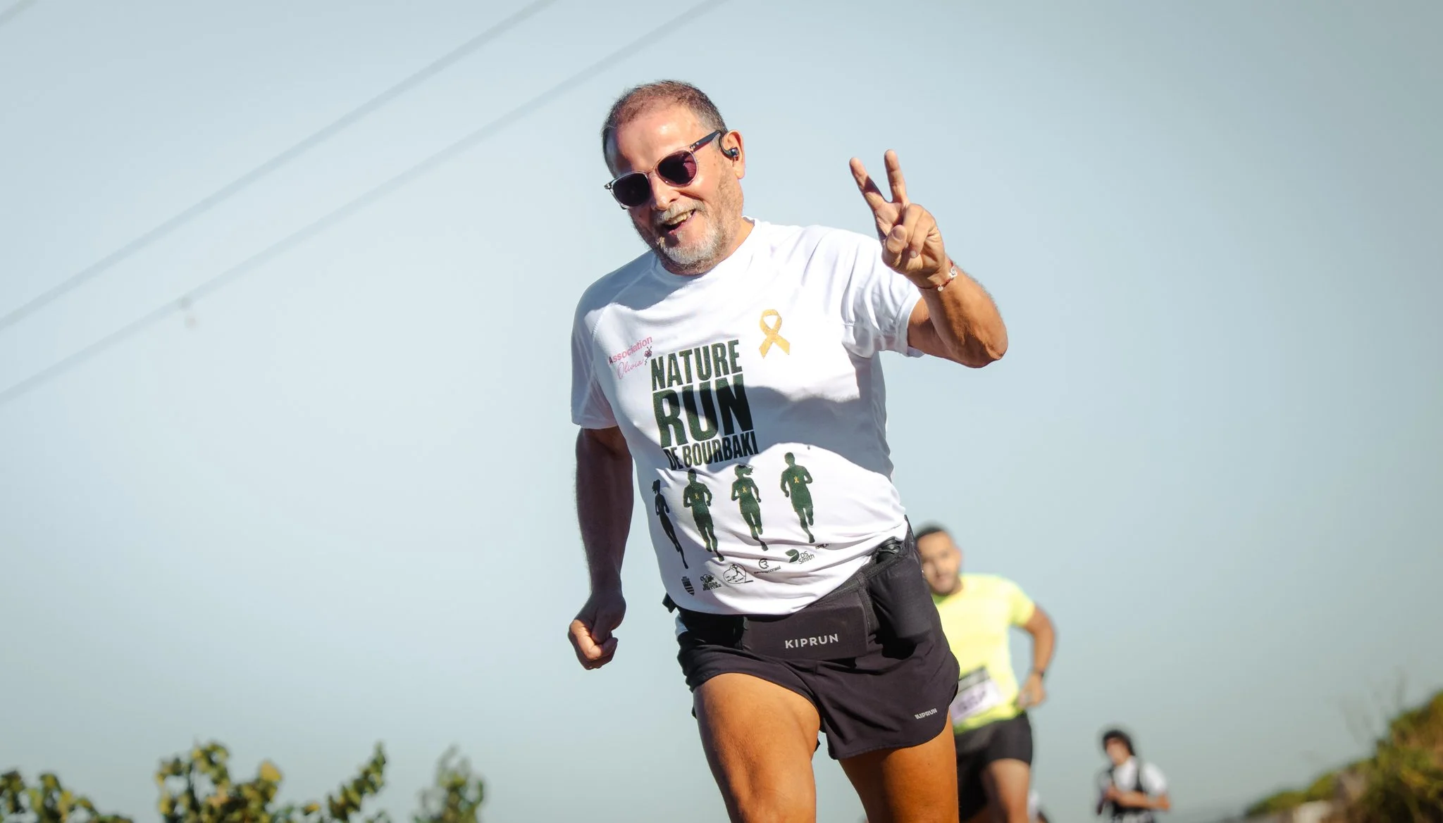 Un homme courant avec un sourire, portant des lunettes de soleil, un t-shirt blanc avec le texte 'NATURE RUN' et une image de coureurs, dans un paysage en plein air avec un ciel clair.
