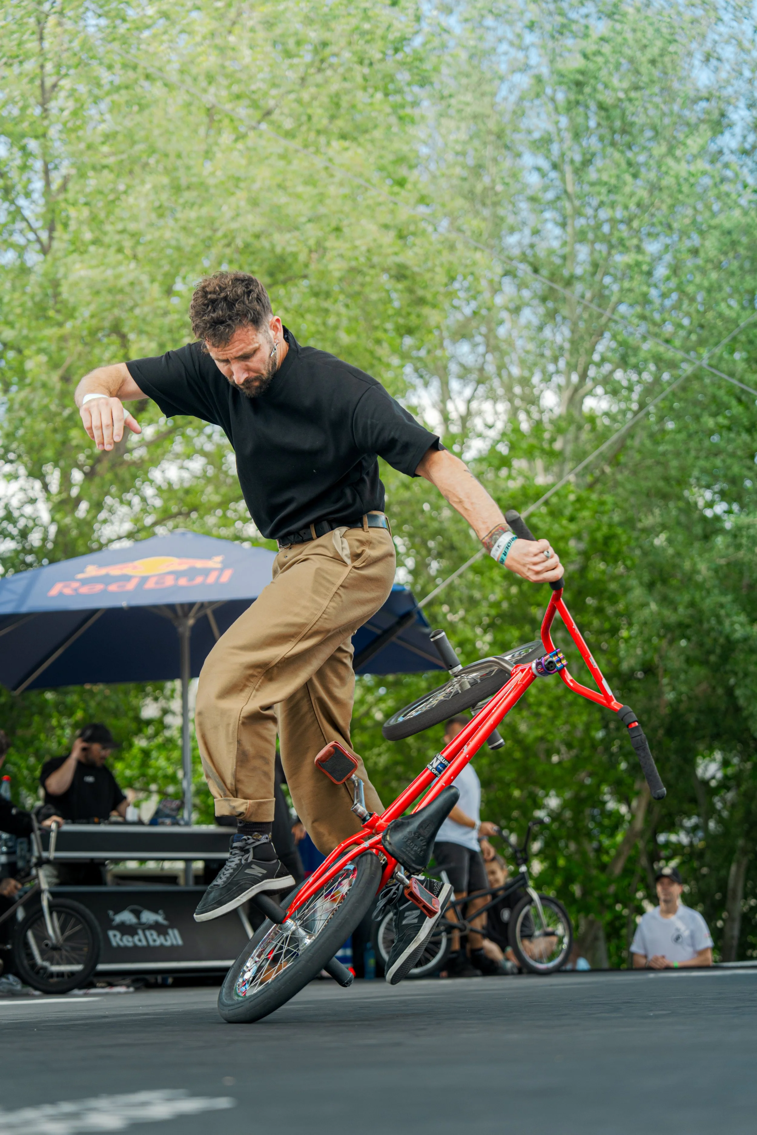 A man performing a stunt on a BMX bike at an outdoor event with a Red Bull sponsor tent in the background, with people and trees surrounding the scene.