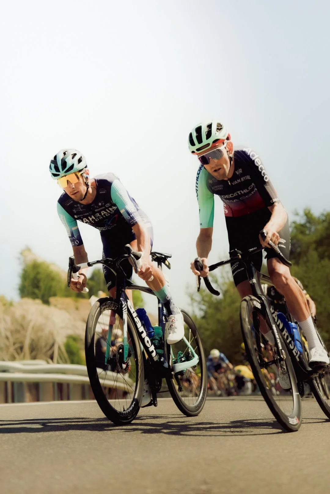 Two cyclists participating in a race, wearing helmets, sunglasses, and cycling jerseys, riding on a paved road with a grassy landscape and trees in the background.