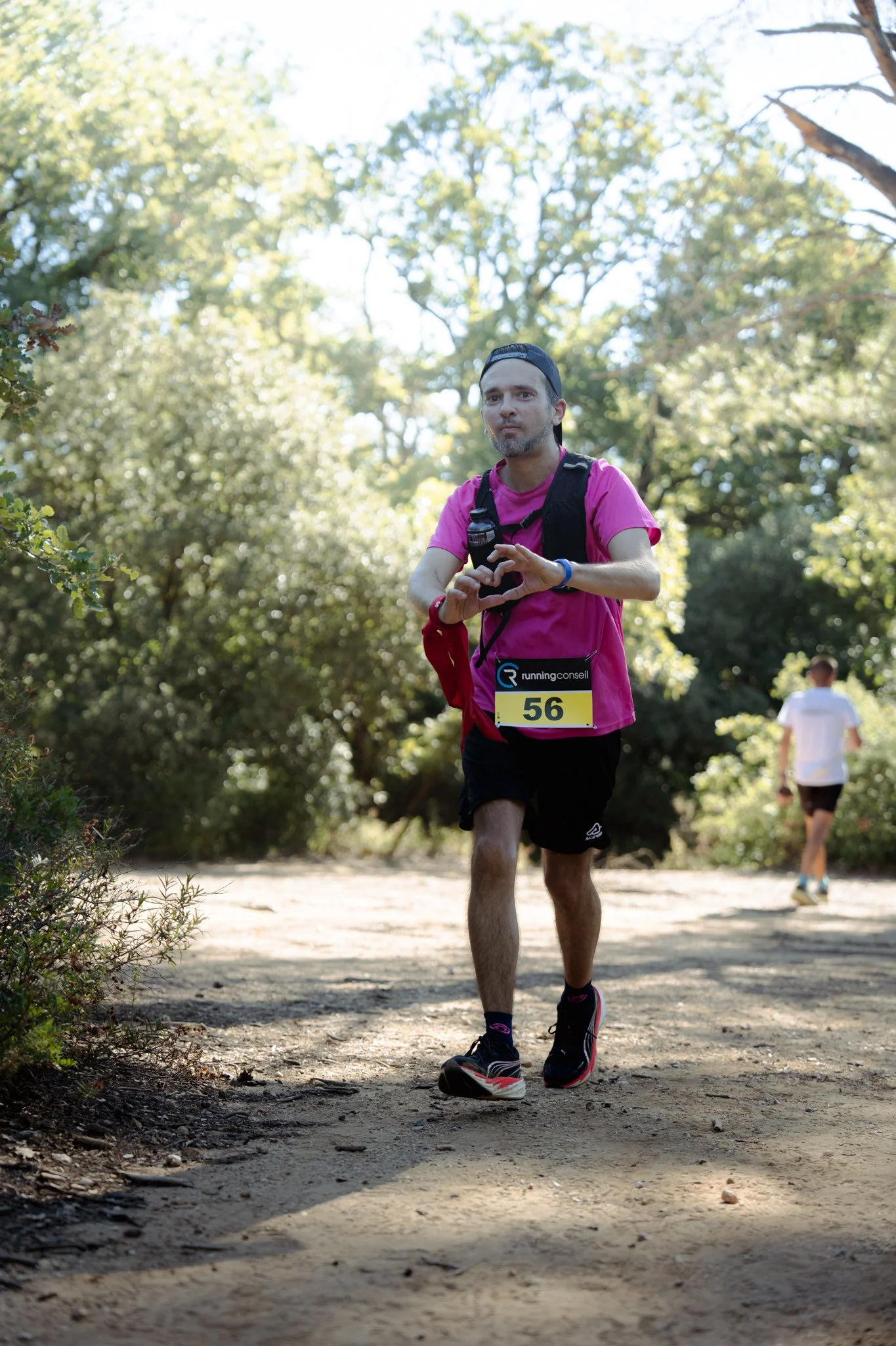 Un homme court dans un sentier forestier, portant un t-shirt rose, un sac à dos, une montre et un dossard numéro 56, faisant partie d'une course ou d'un événement sportif.