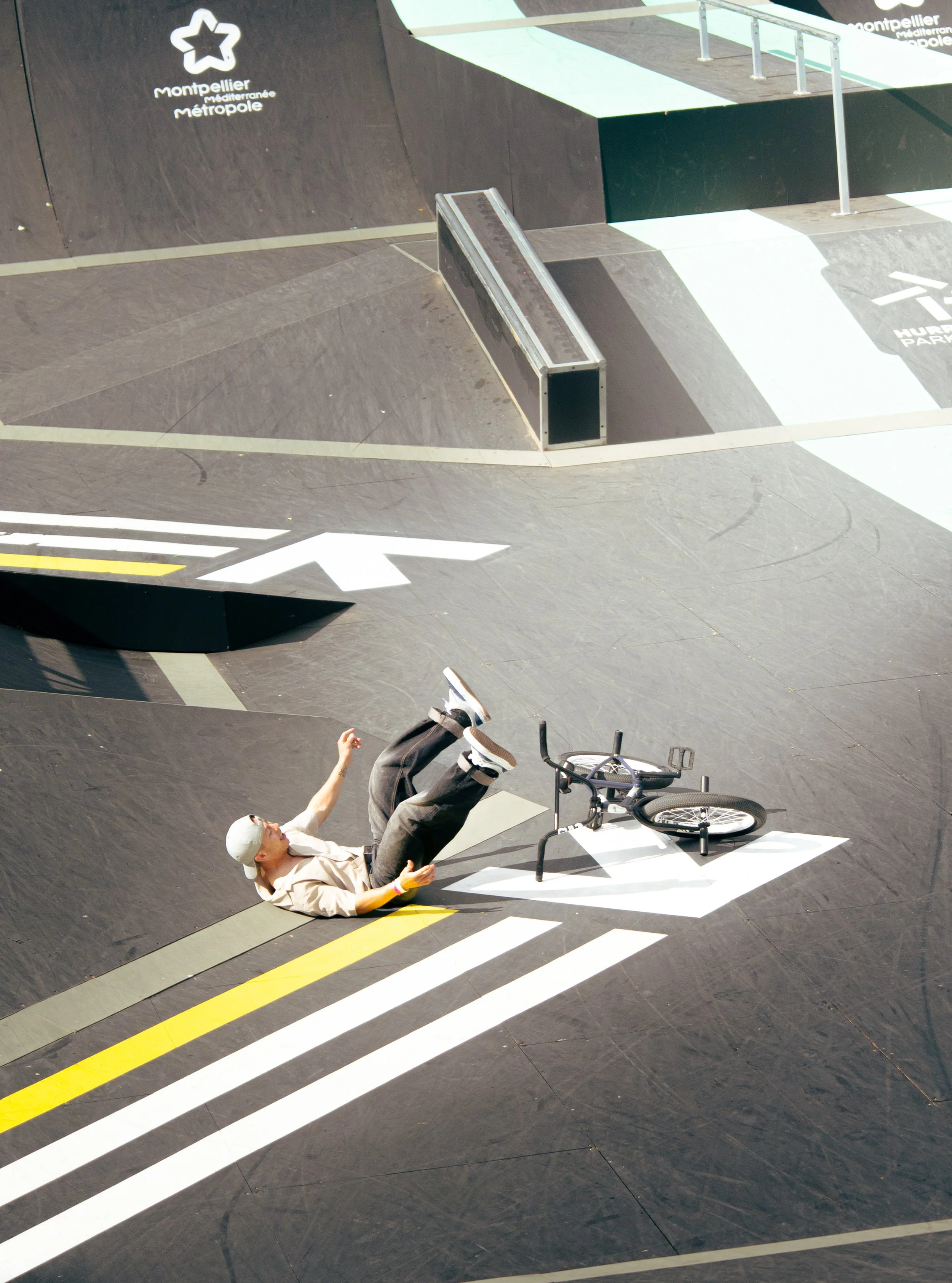 Une personne en skateboard, portant un casque blanc, est tombée sur un skatepark à Montpellier, avec un vélo à côté. La vue est inversée, comme si l'image était prise par un miroir.