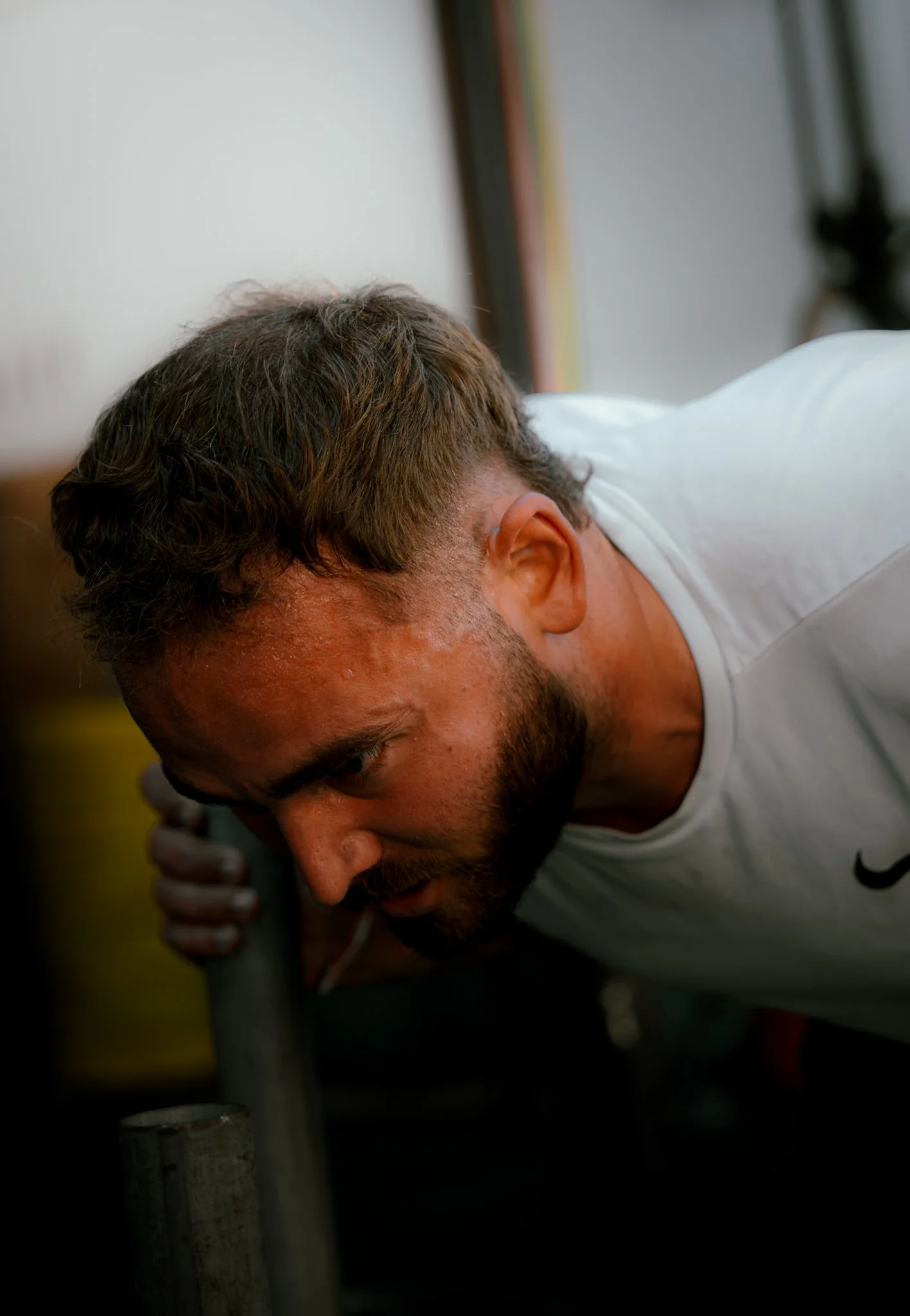 Homme penché en avant, portant un t-shirt blanc, avec des cheveux courts et une barbe, concentré sur une tâche.
