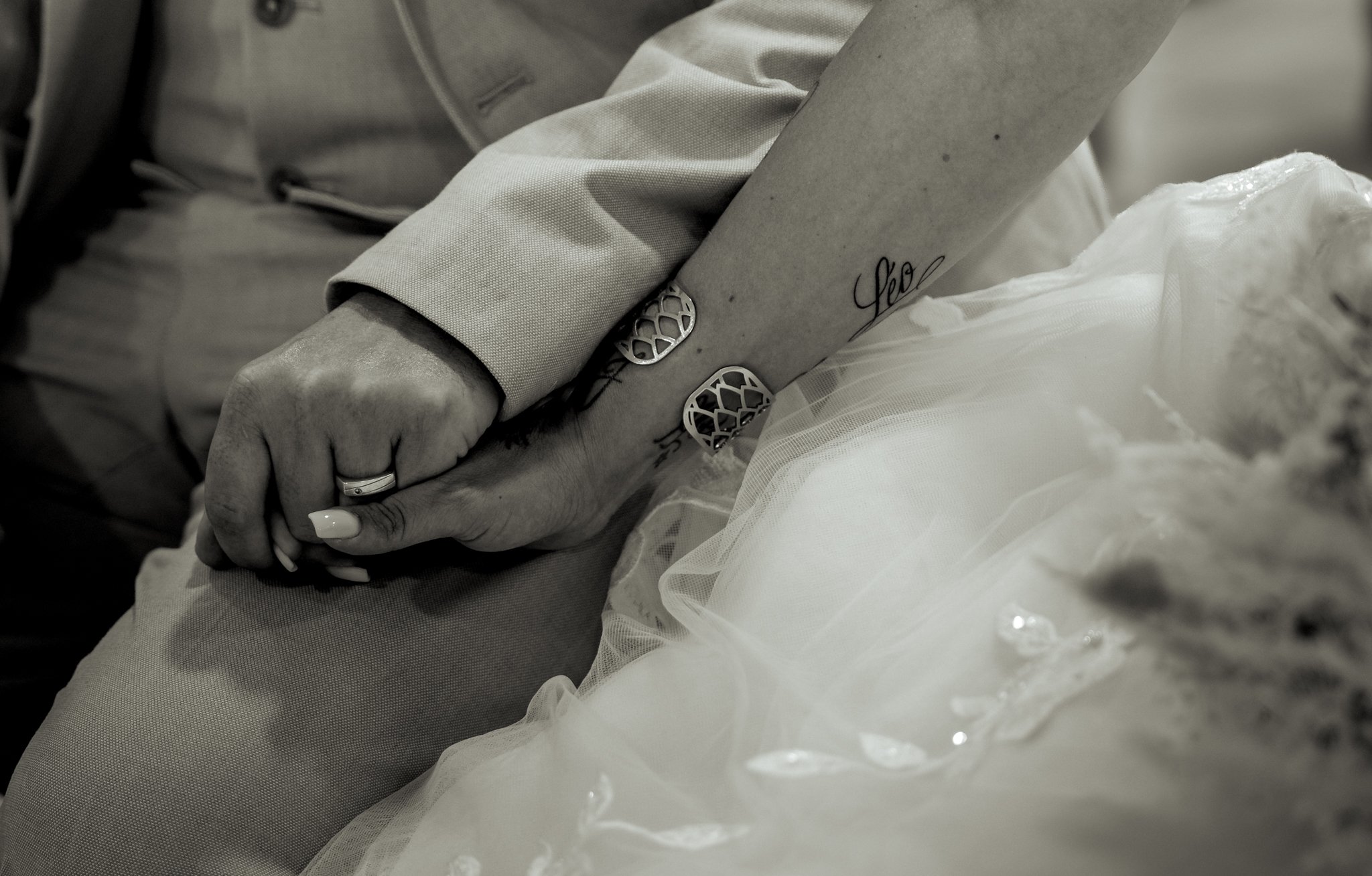 Mains d'une personne portant une bague, tenant une autre main dans un environnement éclairé, habillée en blanc. La scène est en noir et blanc.