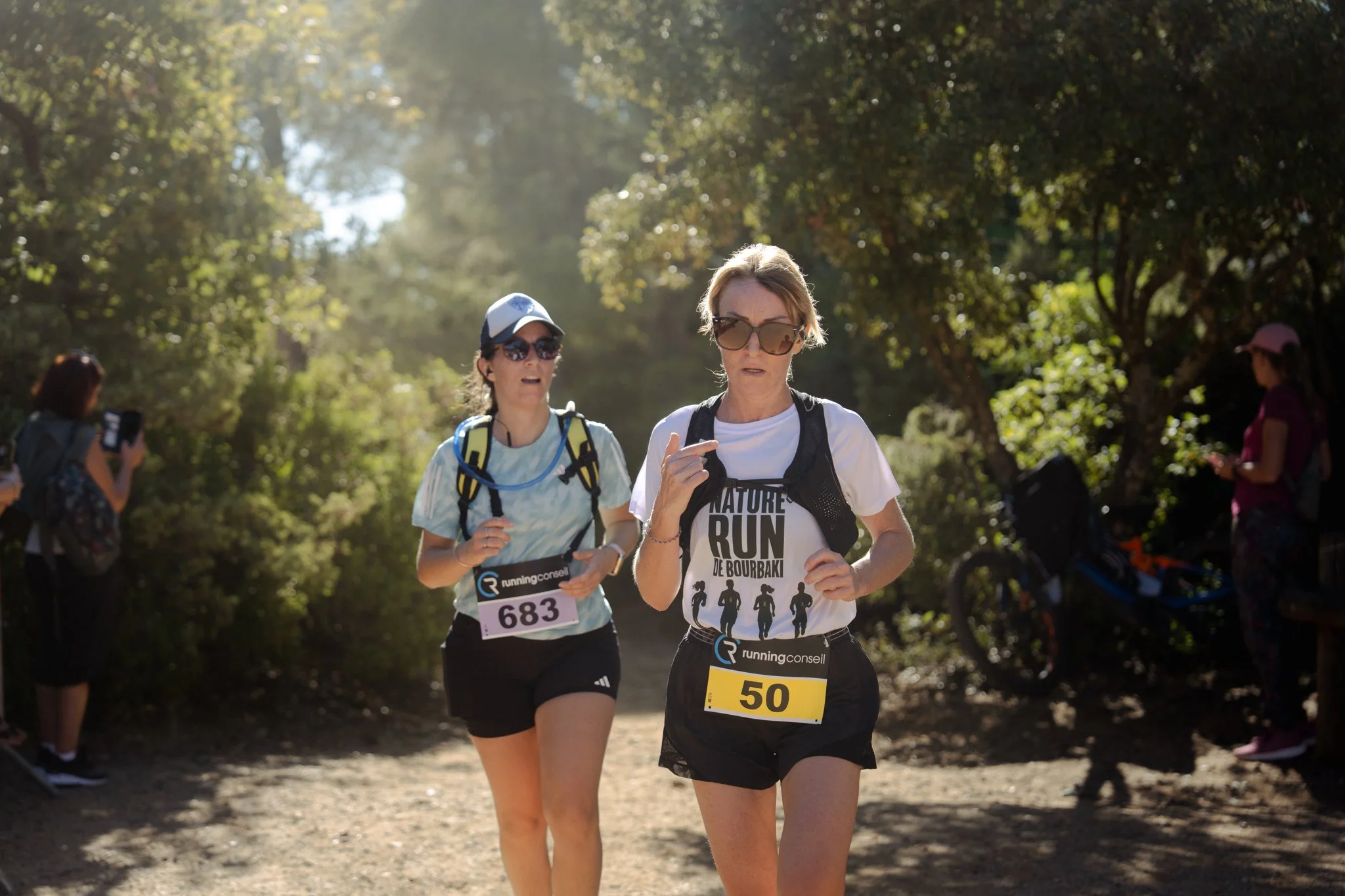 Deux femmes en train de courir lors d'une course en plein air, portant des numéros de course 683 et 50, sous un ciel ensoleillé, entourées d'arbres et de personnes spectatrices.