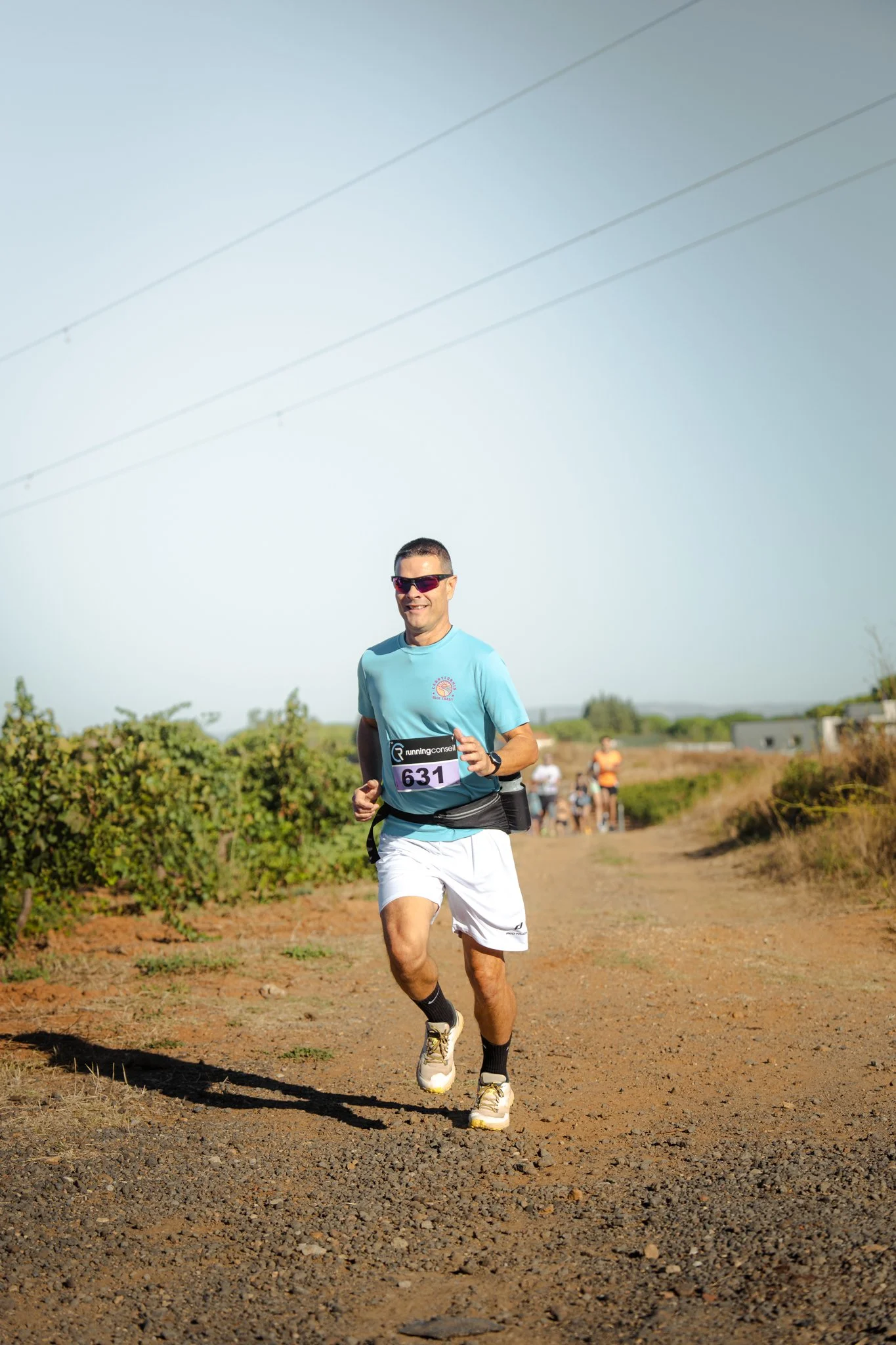 Un homme court sur un chemin de terre pendant une course, portant un t-shirt bleu, un short blanc, une montre, des lunettes de soleil et un numéro de course 631. Il y a d'autres coureurs au fond et le ciel est clair.