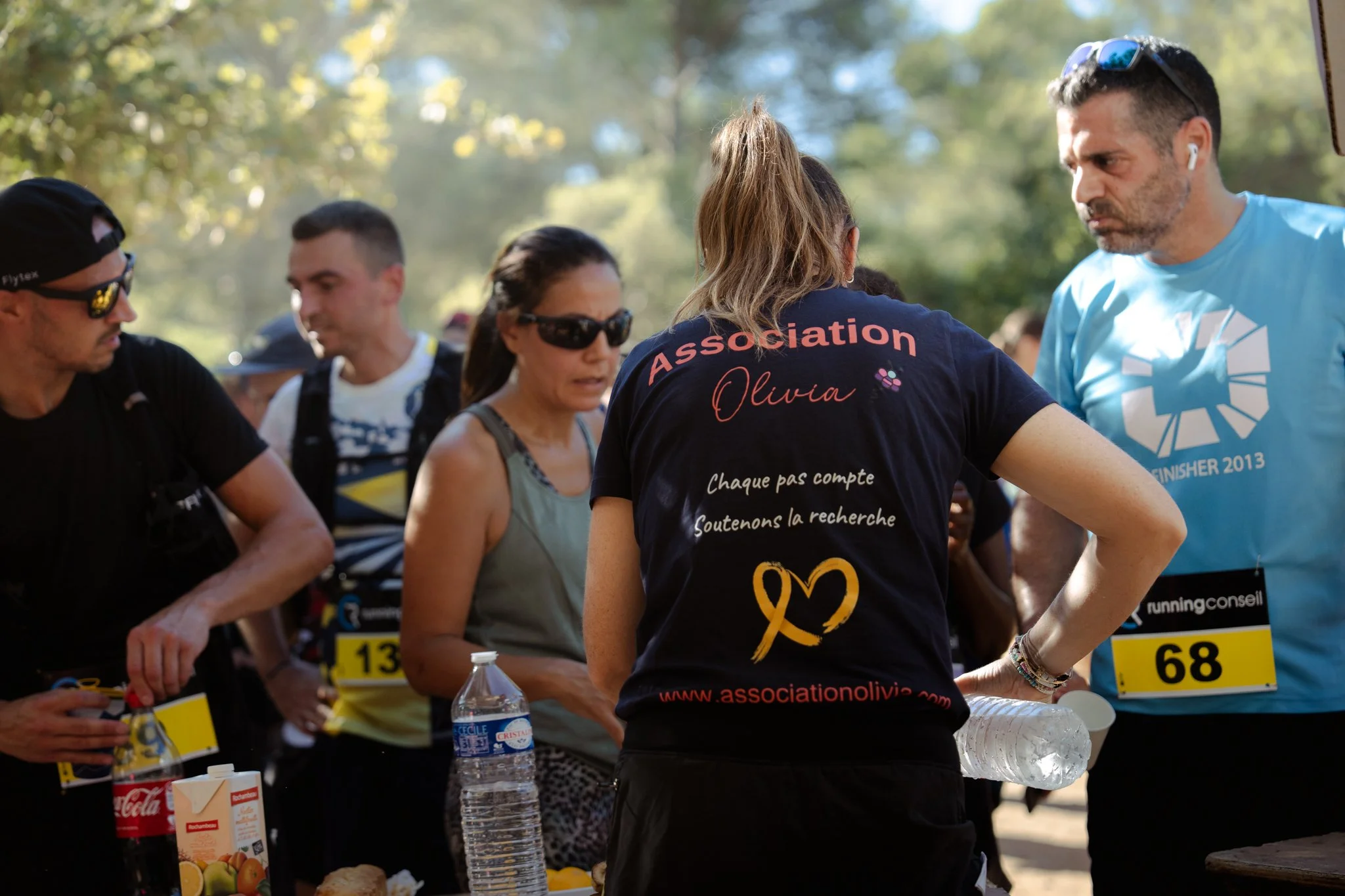 Groupe de personnes participant à un événement de course ou de marche en extérieur, portant des vêtements liés à une association, avec un tableau d'inscription, des bouteilles d'eau, et des numéros de course visibles.