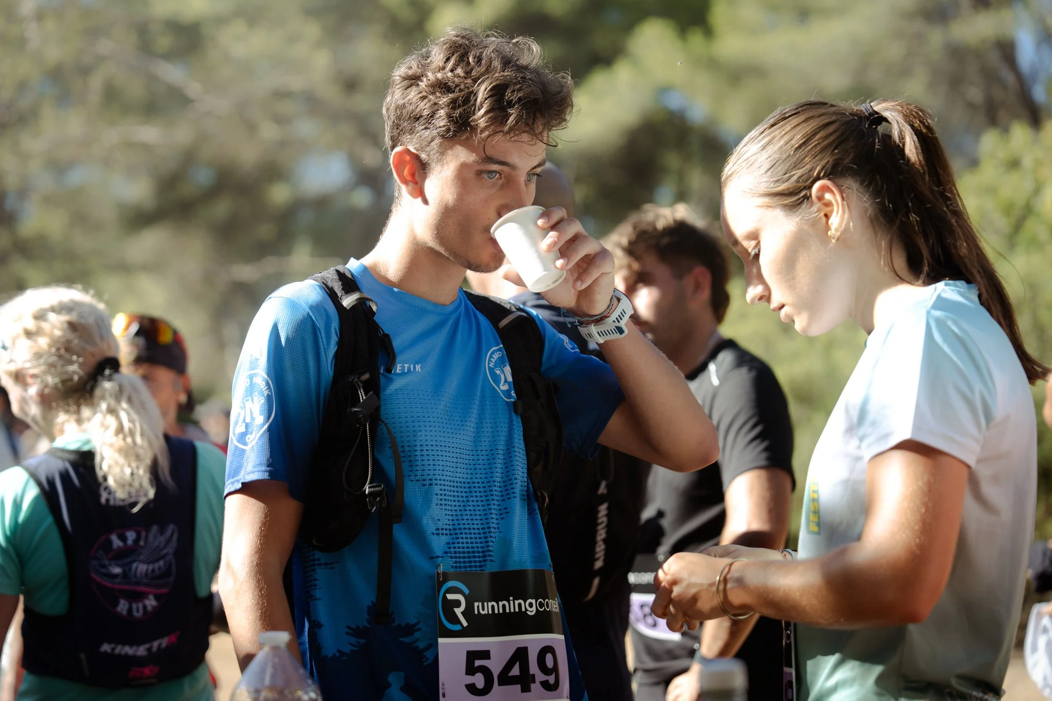 Un groupe de coureurs lors d'une course en plein air, un homme en haut à gauche s'apprête à boire dans une petite tasse, d'autres participants en arrière-plan.
