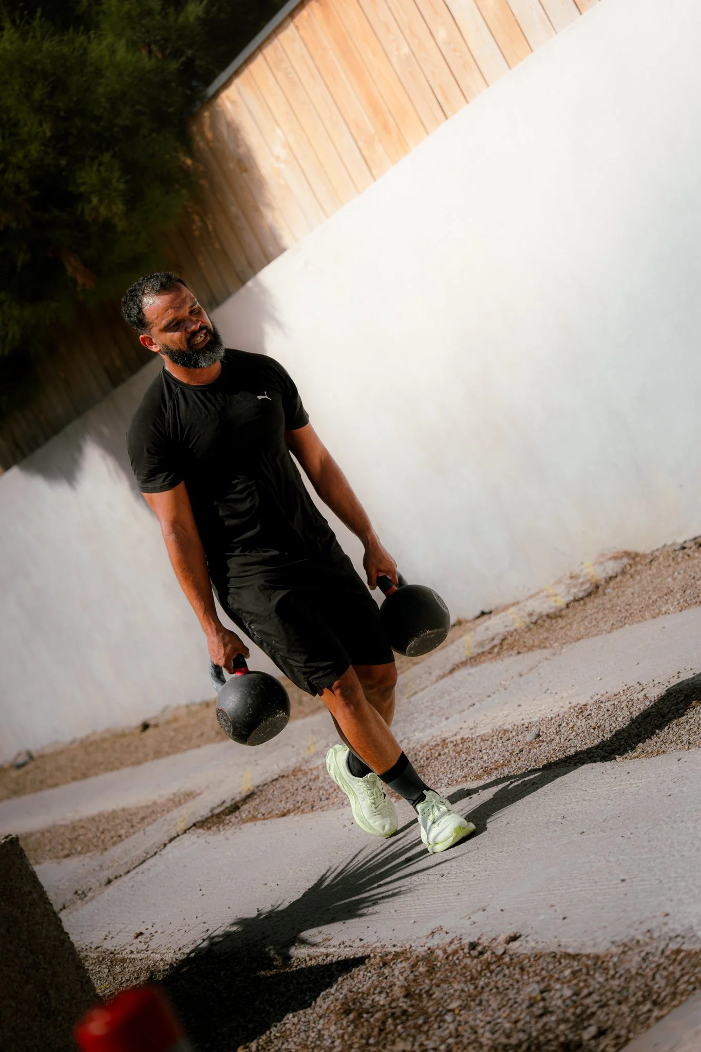 Un homme faisant de l'exercice avec deux haltères en main, portant un t-shirt noir et des chaussures de sport blanches, sur un trottoir en extérieur.